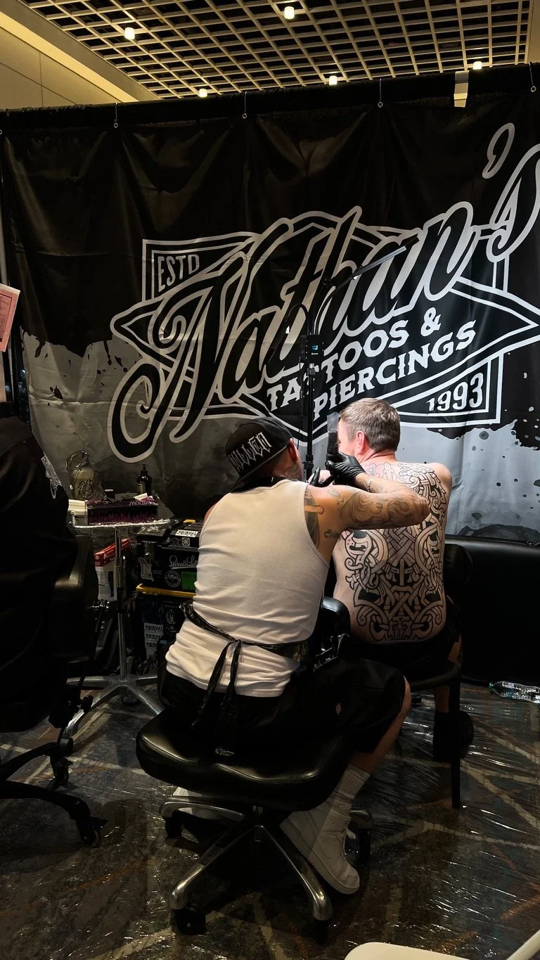 Tattoo artist creates a sleeve tattoo on a man's back at a tattoo convention, with a large black banner displaying the tattoo shop's name in the background.