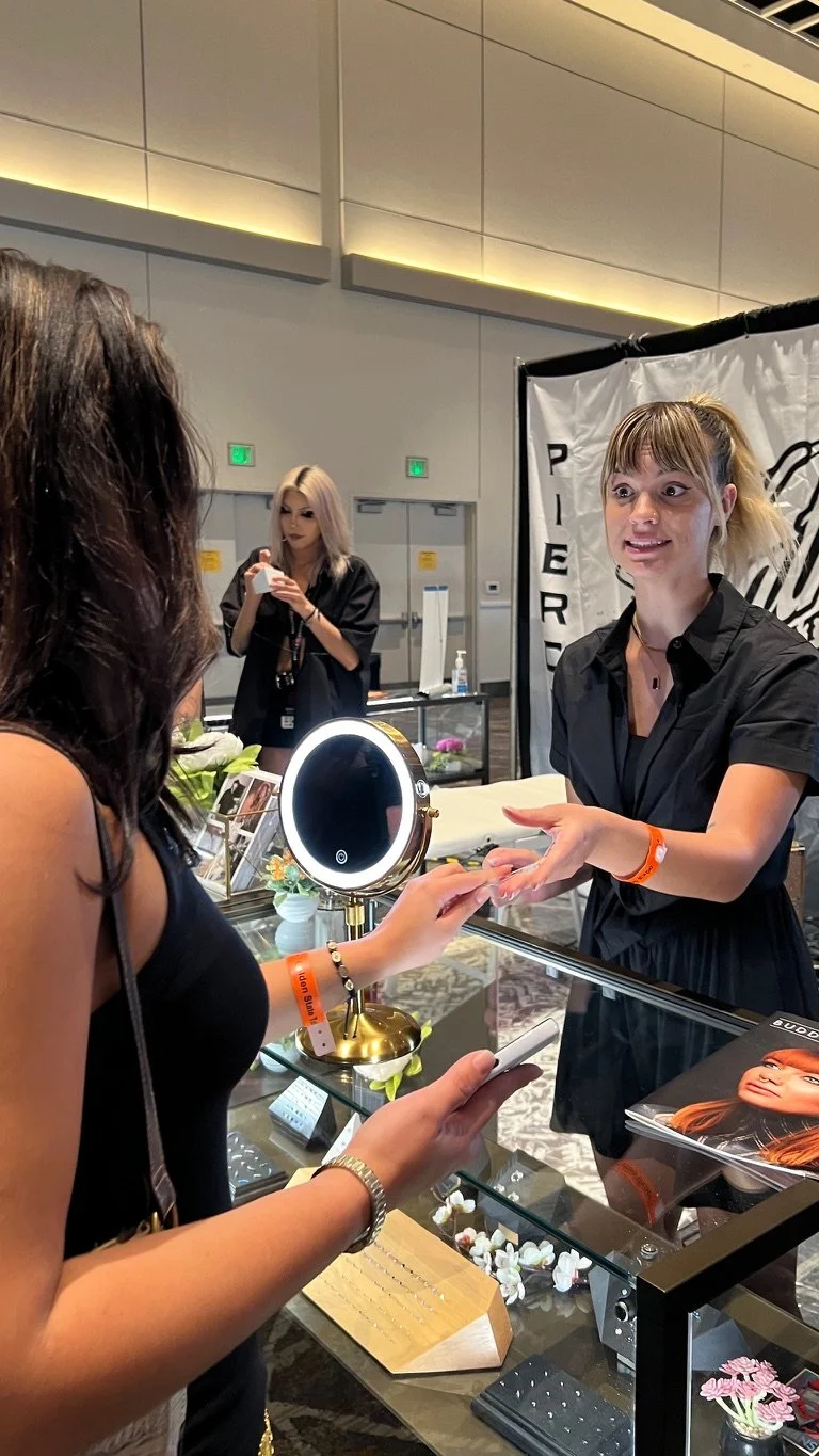 A woman showing jewelry display. The booth features a mirror, flowers, and promotional materials. Another woman in the background is taking a photo.