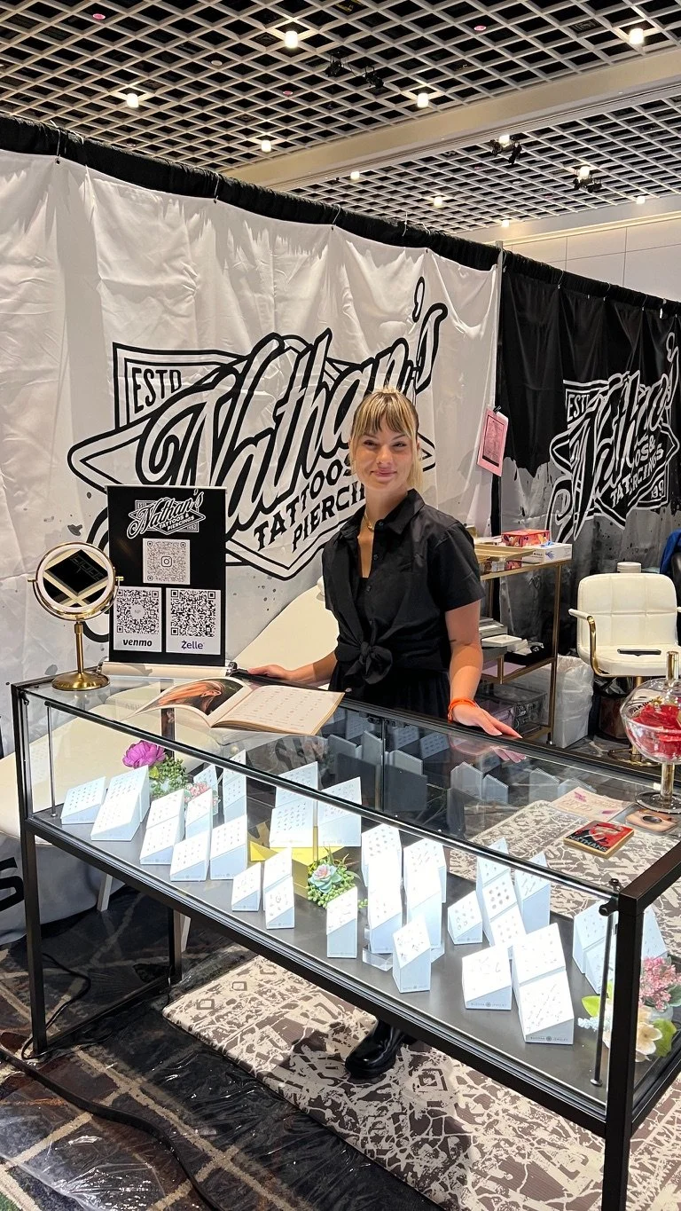 A woman stands behind a glass display counter at a tattoo event, with tattoo designs inside the display case and a sign with QR codes on the counter. She is smiling and wears a black shirt tied at the waist.