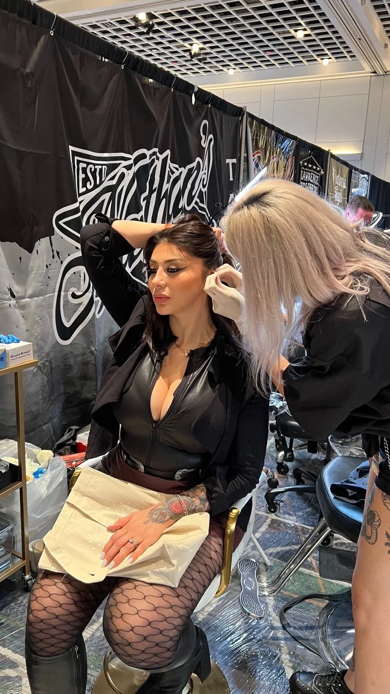 A woman with dark hair, tattoos on her hand and leg, and fishnet stockings is seated in a chair, having her ear pierced by a woman with long blonde hair. The setting appears to be at a convention or event with a black banner in the background and var