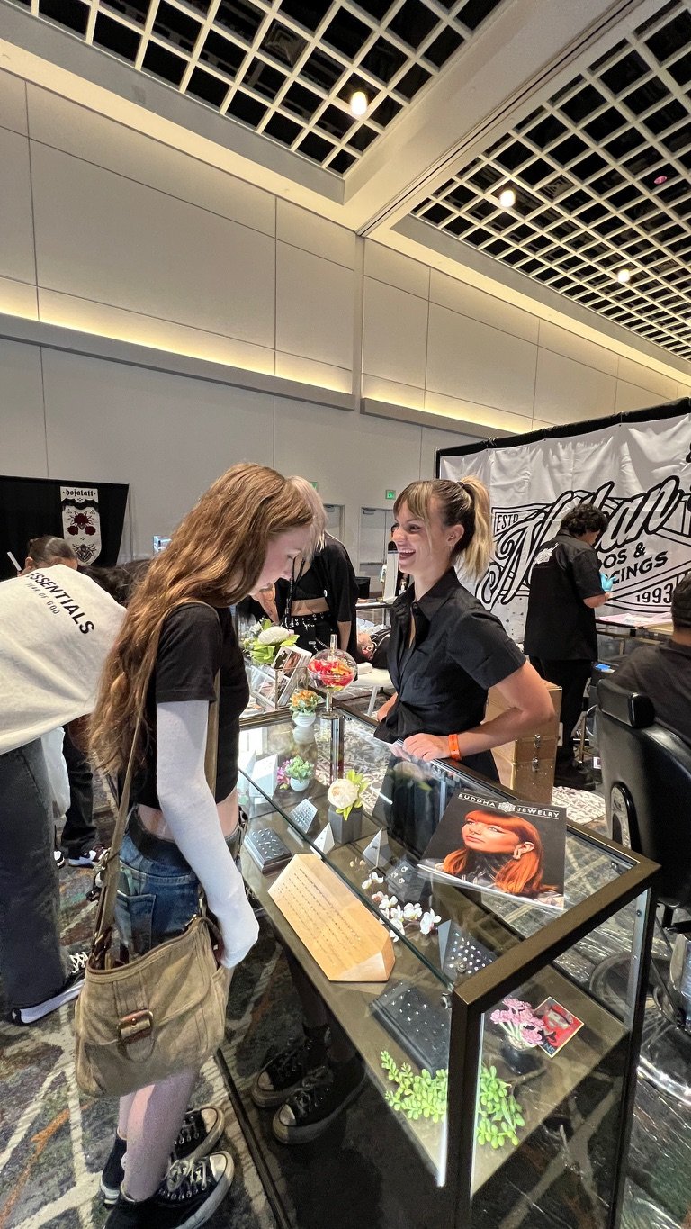 Two women at jewelry booth chatting, with display case of jewelry and plants, behind a black and white banner.
