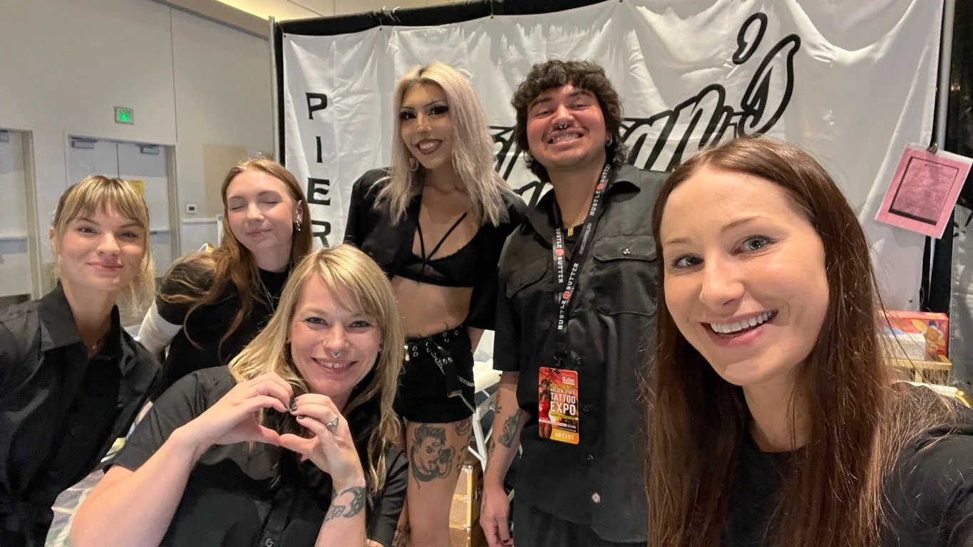 Group of six people, five women and one man, taking a selfie at a tattoo expo, standing in front of a banner with the event's logo.