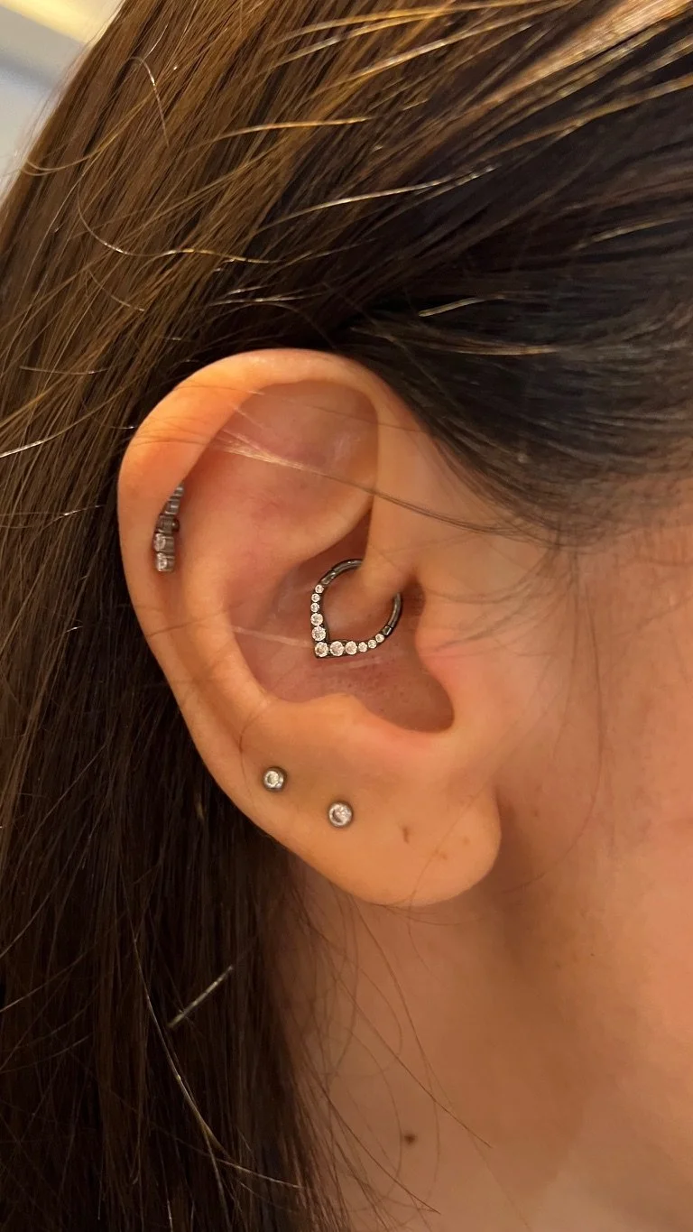 A close-up of a person's ear with multiple earrings: a small hoop with clear stones, a half-circle hoop with clear stones, and two small stud earrings with clear stones.