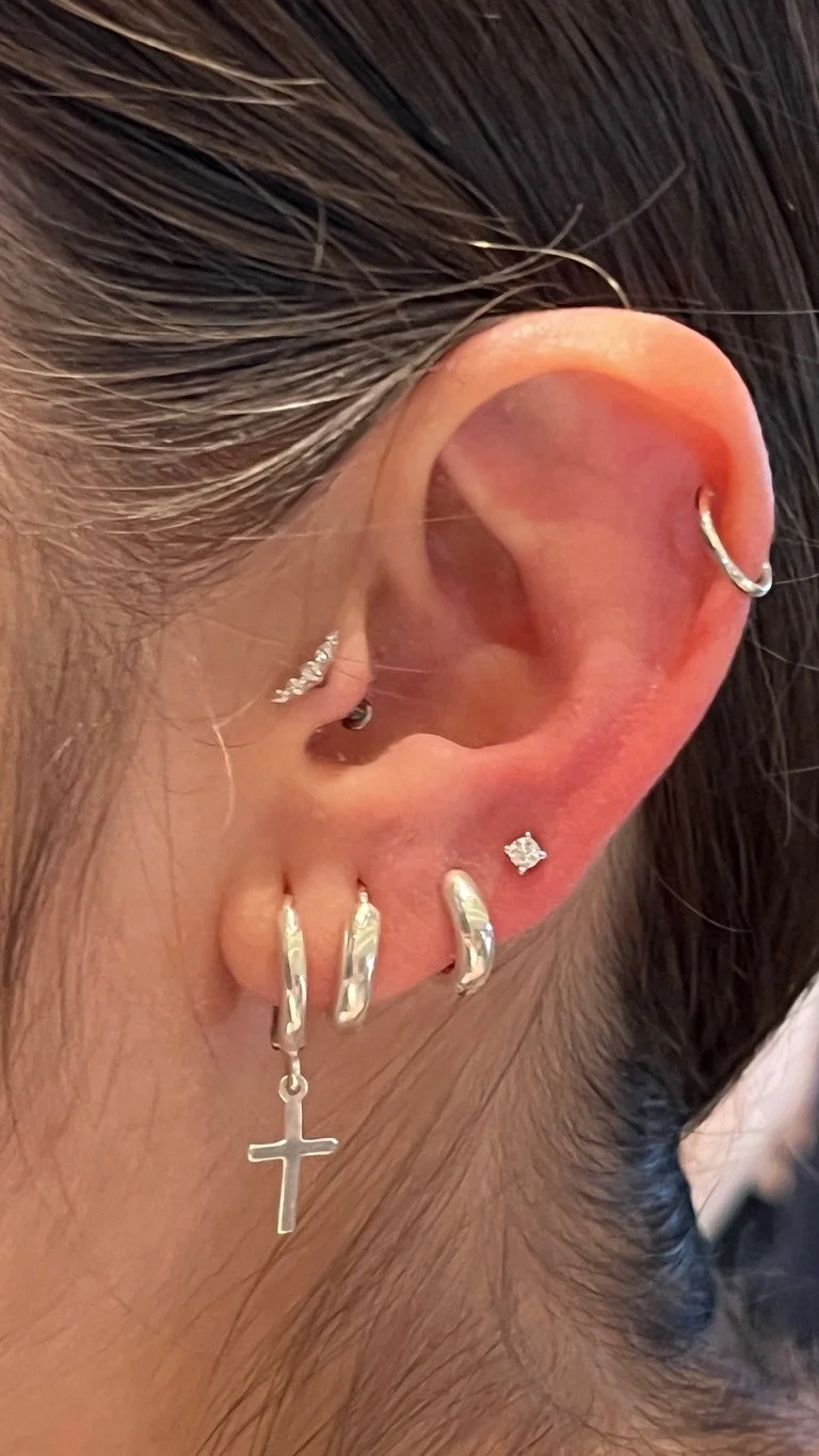 Close-up of a person's ear with multiple earrings, including a hoop with a cross charm, a small stone earring, and several other hoops.