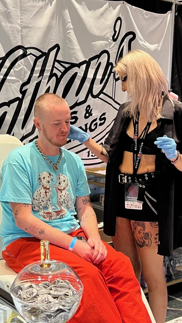 A man with tattoos and a chain necklace sitting with his eyes closed as a woman with blond hair, tattoos, and wearing gloves, touches his head. They are at a convention or event, with a large sign behind them and a glass container with a straw in the