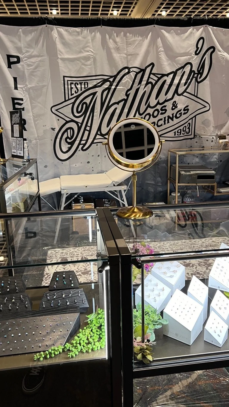 Jewelry display case with rings and earrings, a small decorative plant, and a mirror, in front of a backdrop with the logo 'Athens' and the words 'Pier 9 Toys & Piercings'.