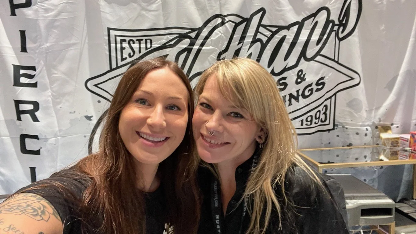 Two women smiling and posing together at an event, standing in front of a large banner with the words 'Harley Davidson' and 'Est. 1993' printed on it.