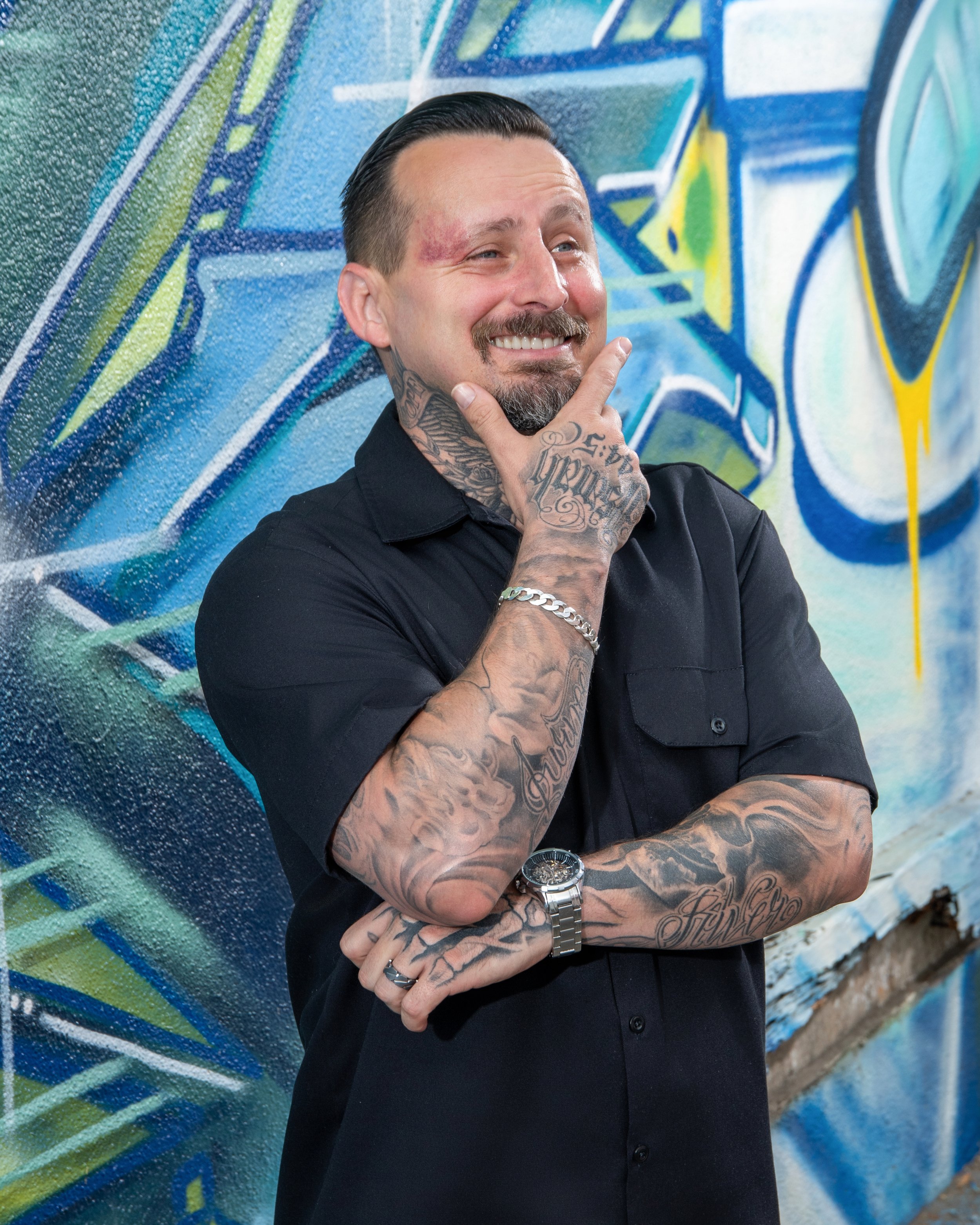 A man with tattoos and a mustache, smiling and posing with his hand on his chin, standing in front of a colorful graffiti wall.