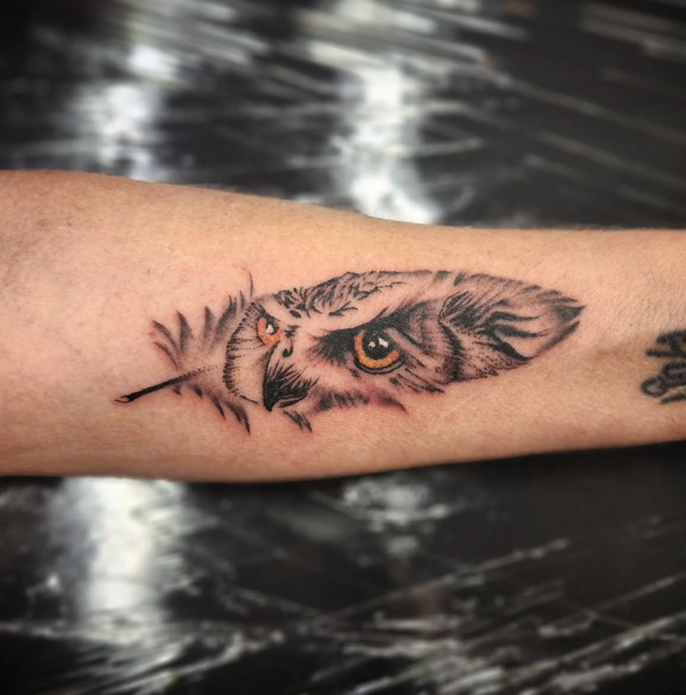Close-up photo of a tattoo on a person's skin, featuring a detailed tiger's face with orange eyes, set against a blurred background of metallic and shadowy textures.