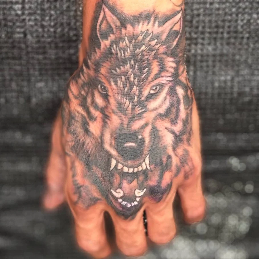 Tattoo of a fierce wolf's head with bared teeth on a person's hand.