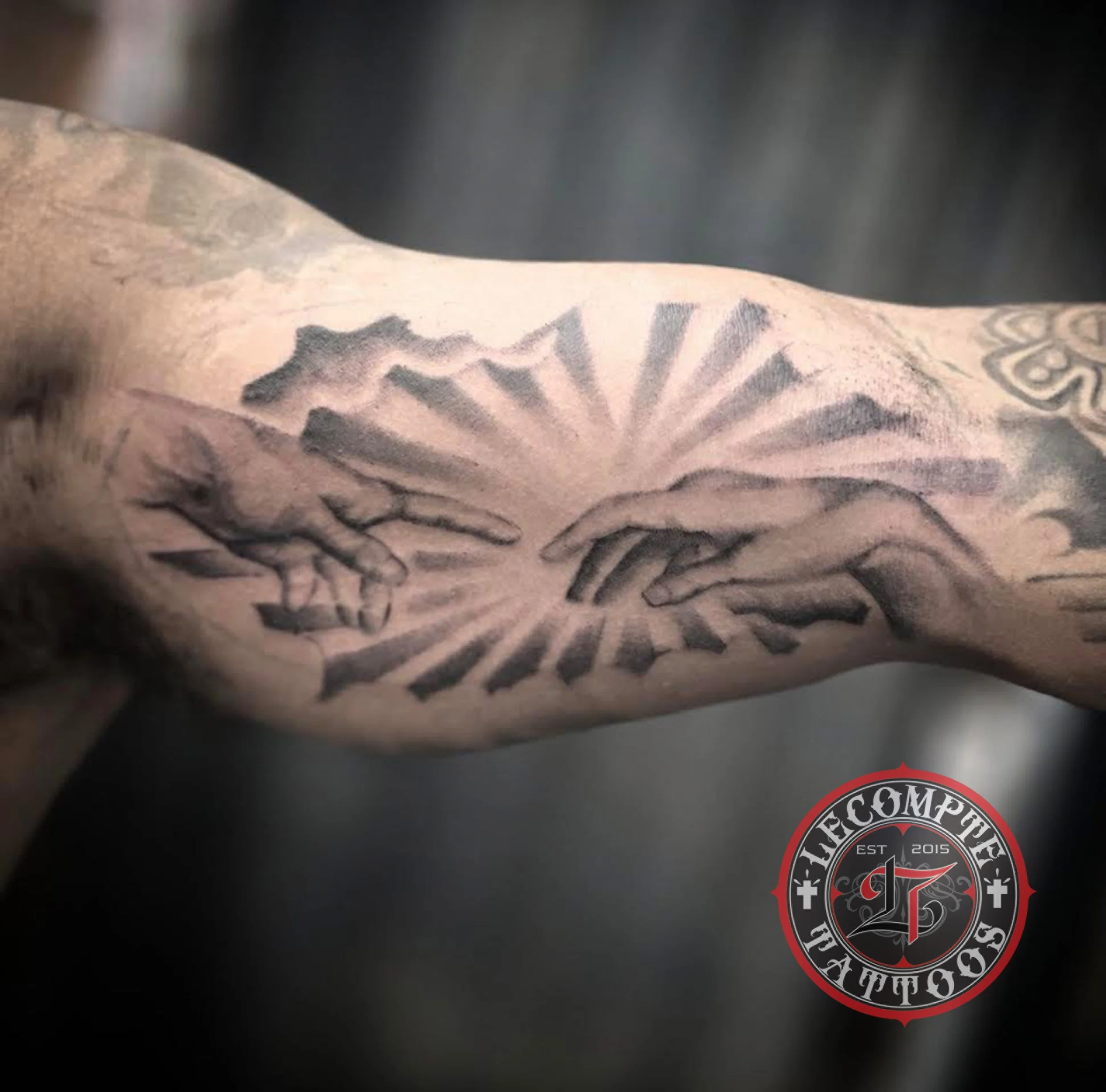 Close-up of a tattooed arm with a prominent black and gray tattoo, slight ink smudges, and a partial logo or patch with the text 'The Complete (est. 2015)', a cross, and a stylized 'S' at the bottom.
