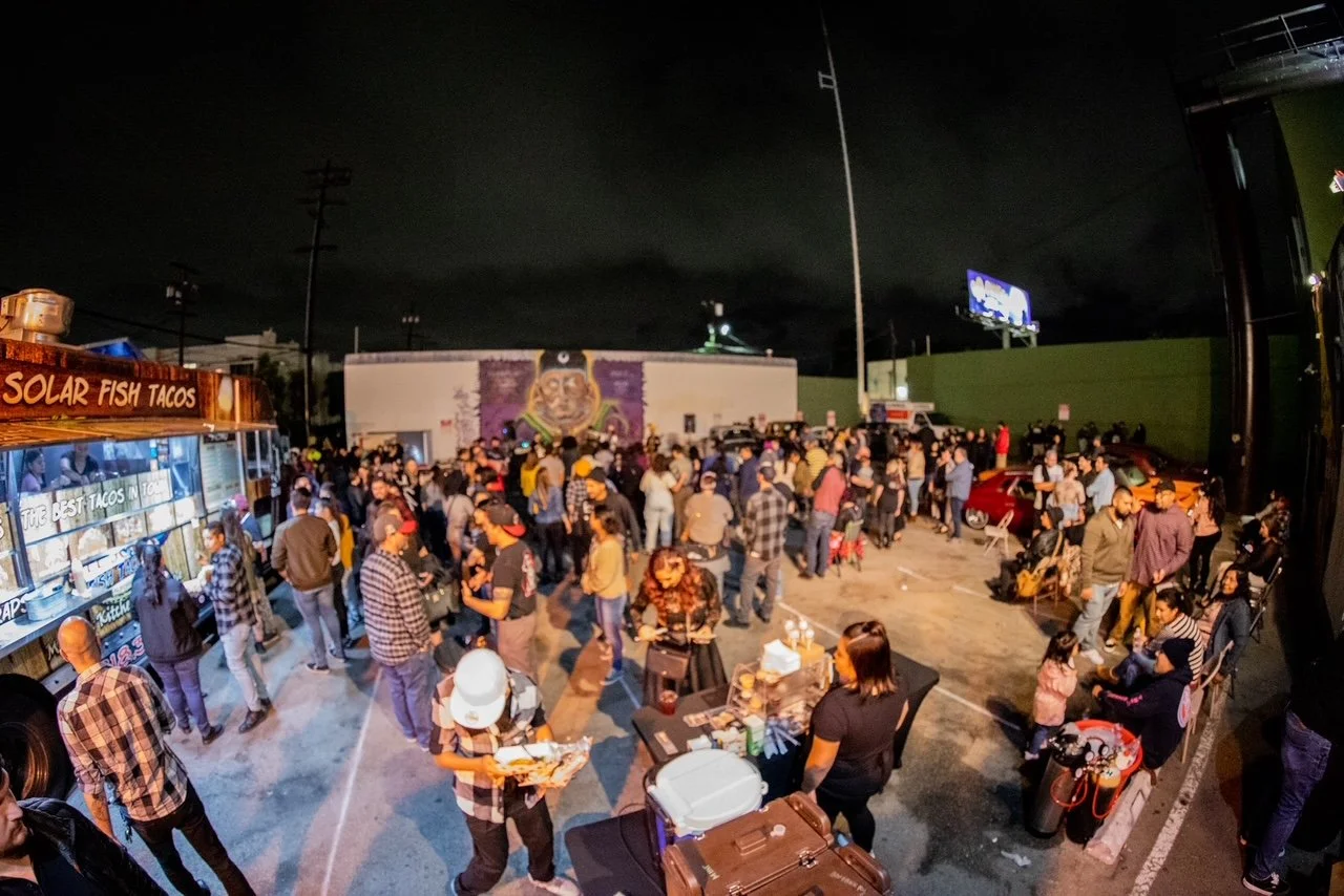 Night scene at a crowded outdoor event with people gathered around a food truck selling solar fish tacos, in a parking lot with cars and a mural on the wall featuring a colorful face.