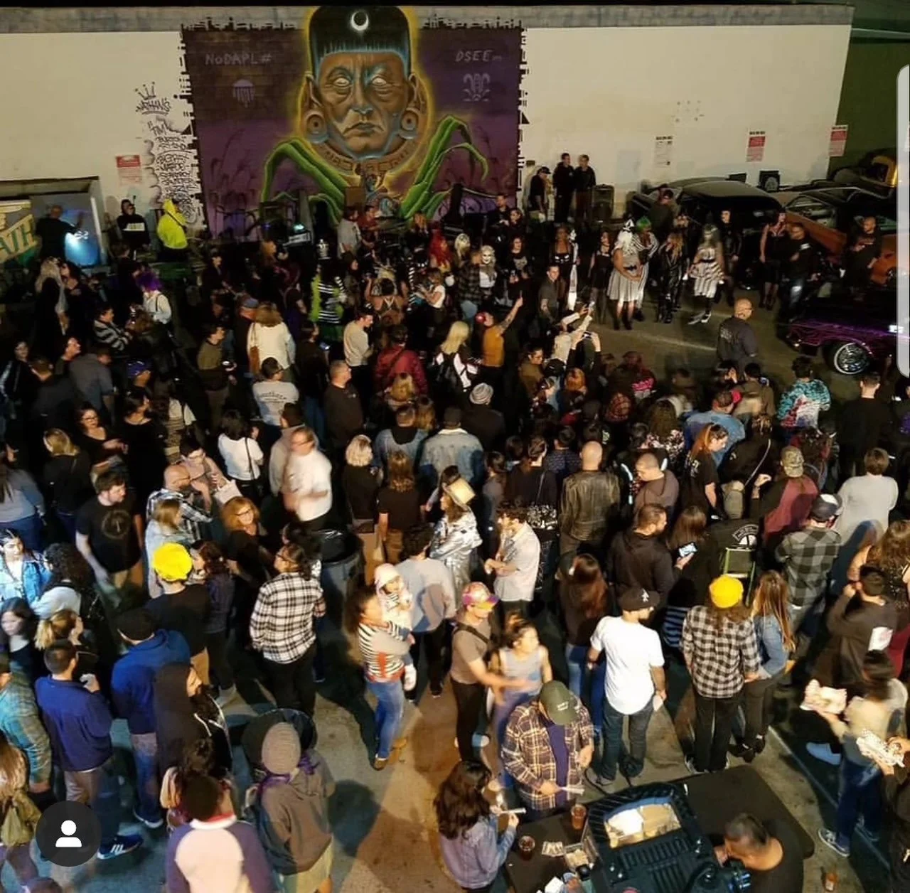 Crowd of people gathered at an indoor event, with some dressed in costumes. There is a large mural of an indigenous person on the wall and vintage cars parked along the sides.