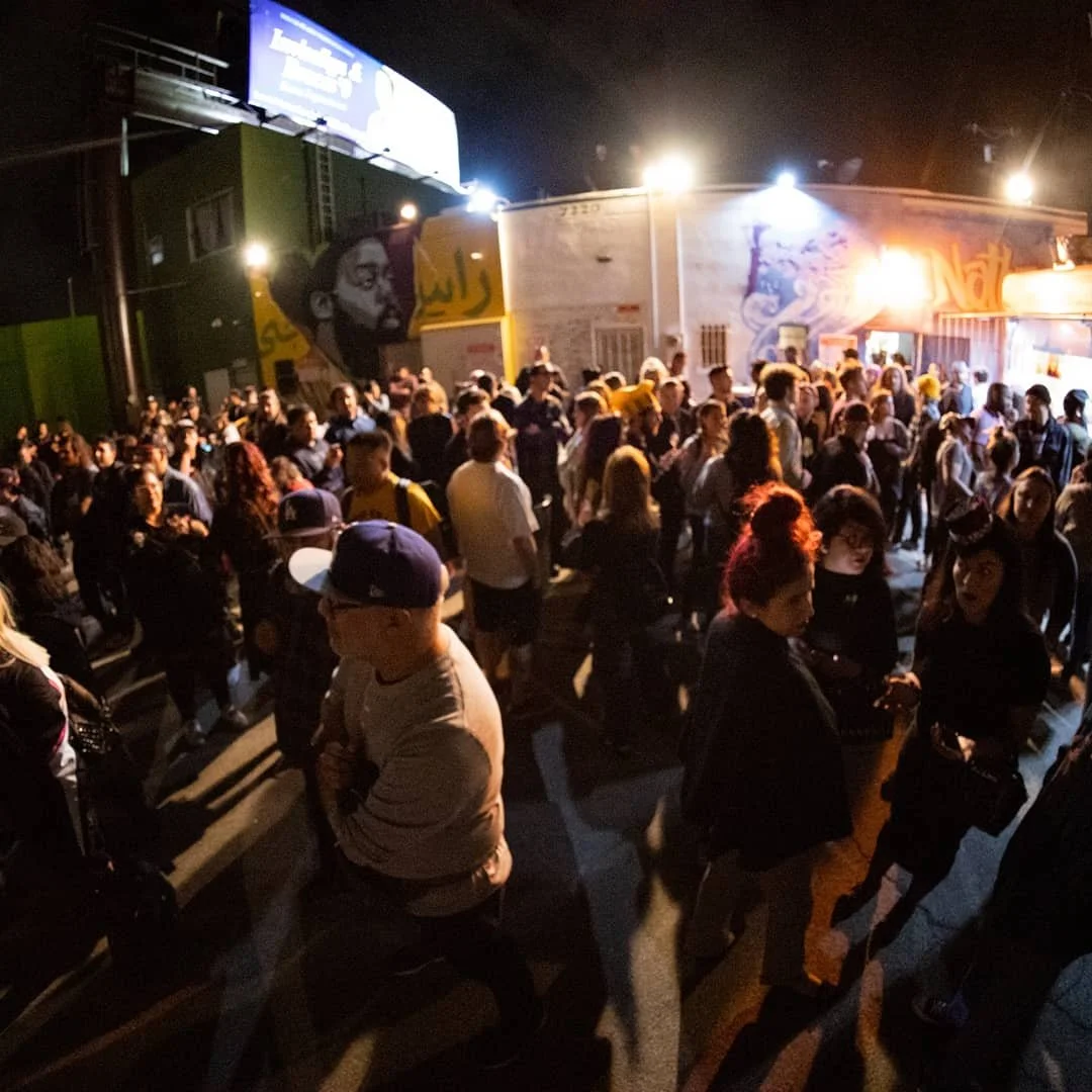 Crowd of people gathered outside at night, some engaged in conversations, with bright lights illuminating the scene. In the background, there is a wall with murals and a billboard.