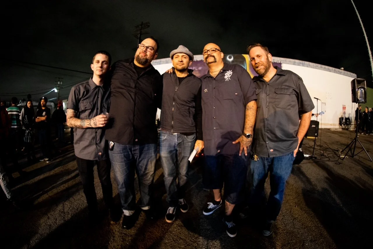 Group of five men posing together outdoors at night, standing on a street with a stage and crowd in the background.