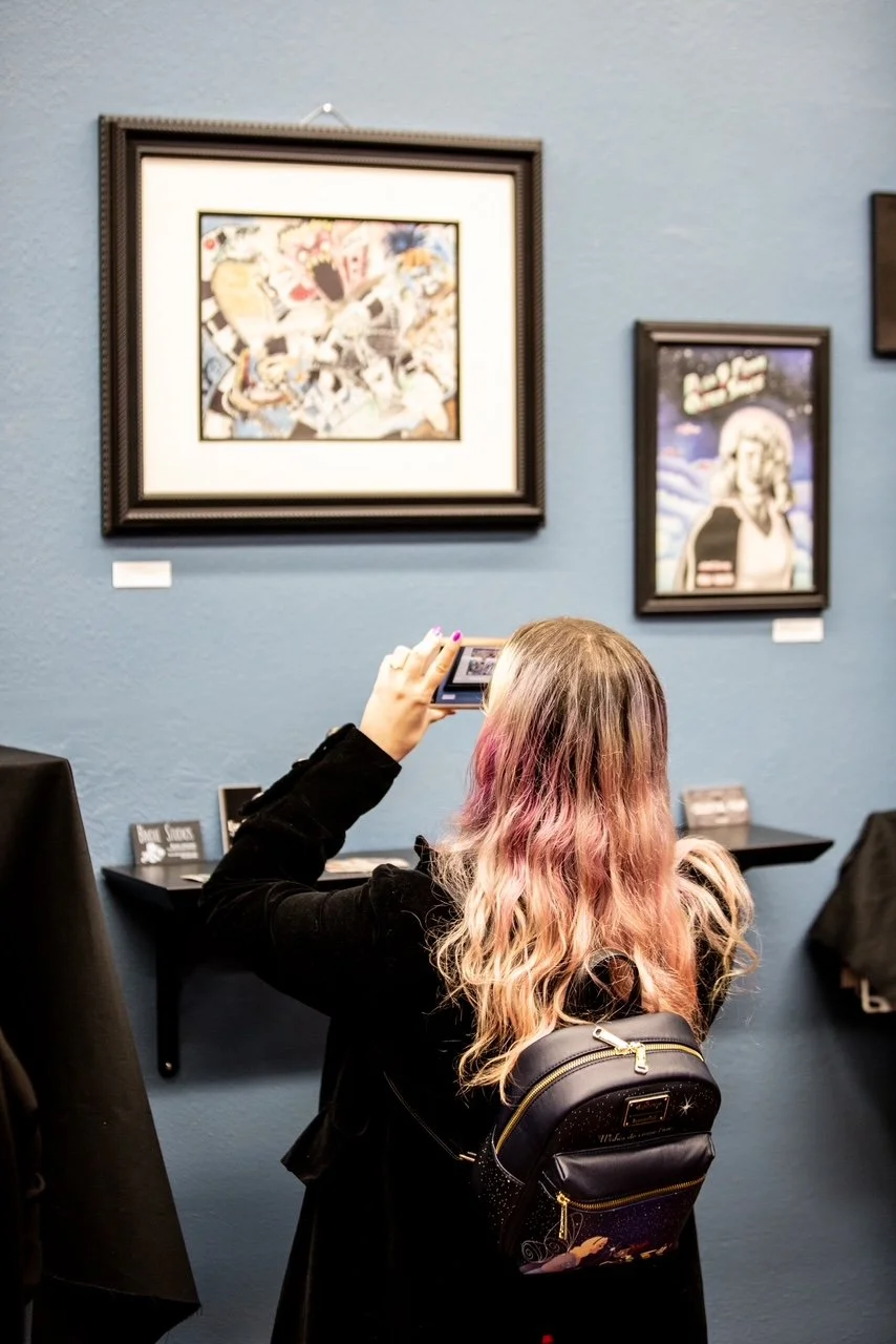 A young woman with pink and blonde wavy hair taking a photo with her phone inside an art gallery, with framed artworks on a blue wall behind her.
