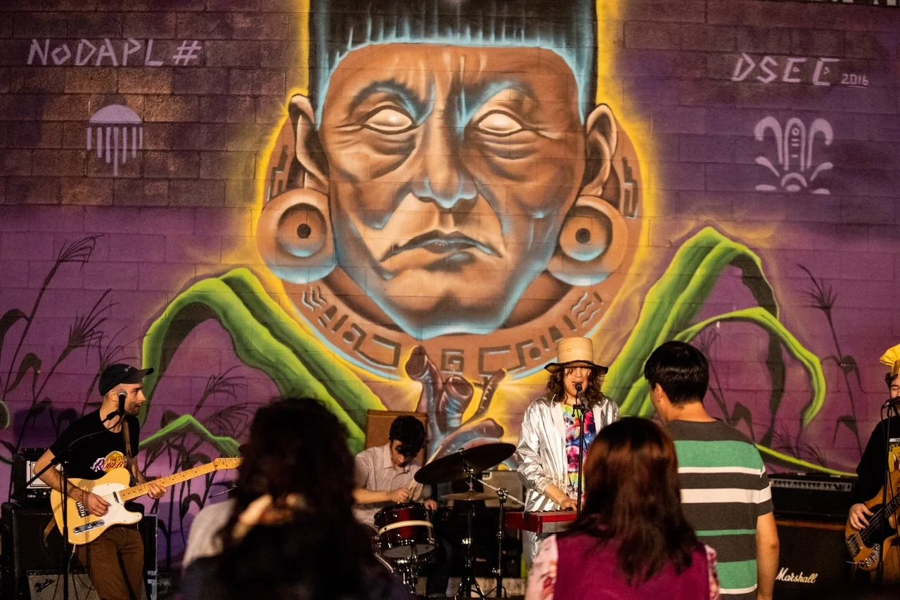 A band performing live in front of a large vibrant mural of an indigenous person with large earrings and traditional garb painted on a brick wall. The mural features purple and green hues with plant motifs in the background.