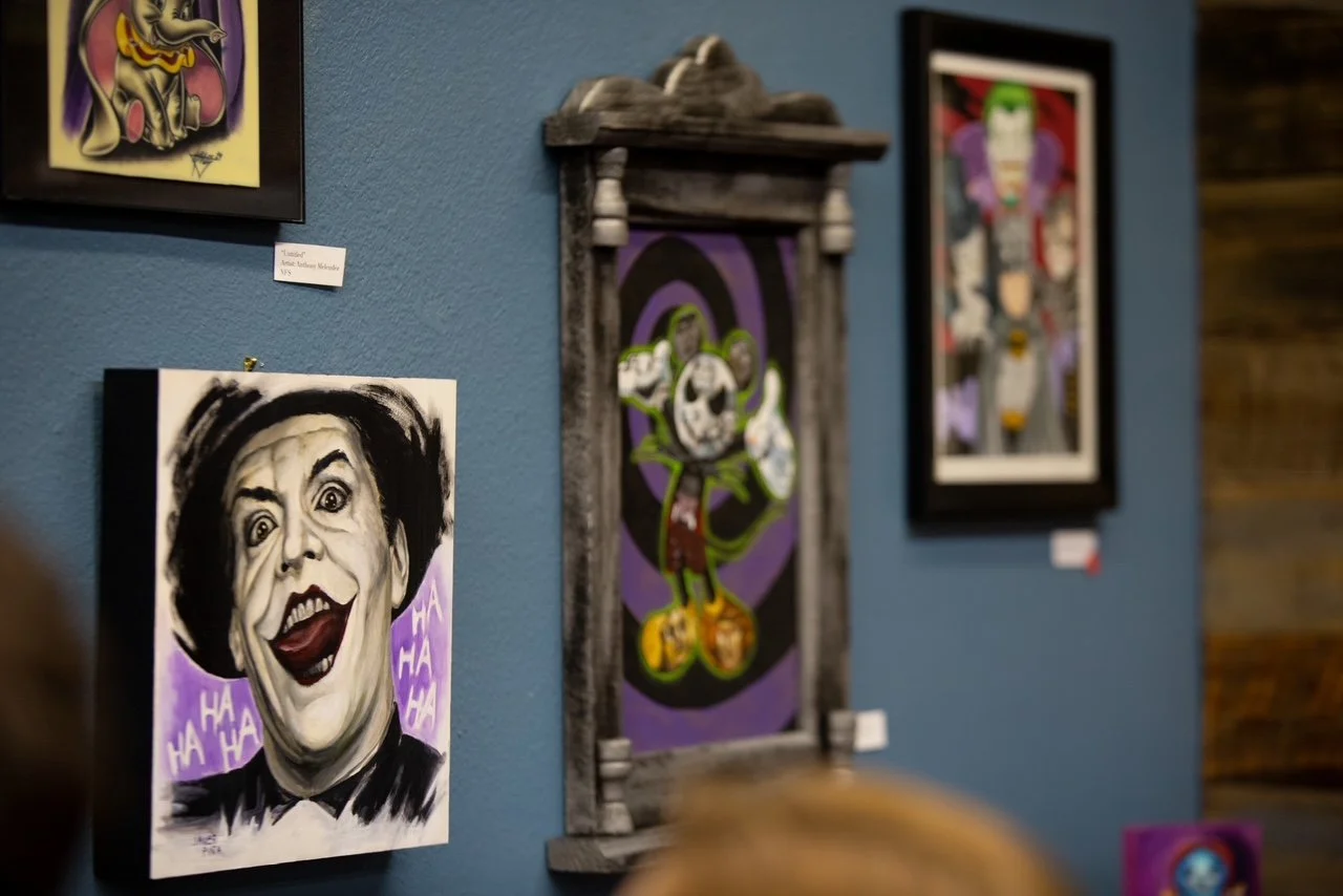 Art gallery display with various colorful portraits and portraits, including a black-and-white portrait of a woman with exaggerated facial features and laughter, and a framed art piece with a character with green hair and various animated features.