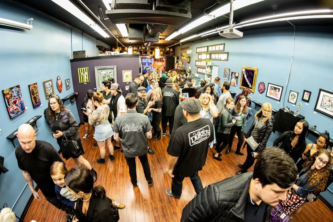A crowded art gallery with people socializing and viewing colorful artwork on blue walls, with a wooden floor and various framed pieces displayed.