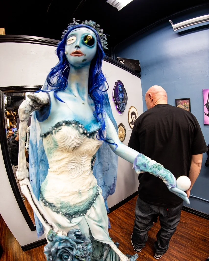 A surreal, artistic sculpture of a female figure with blue skin, long blue hair, and unusual facial features, standing in an art gallery.
