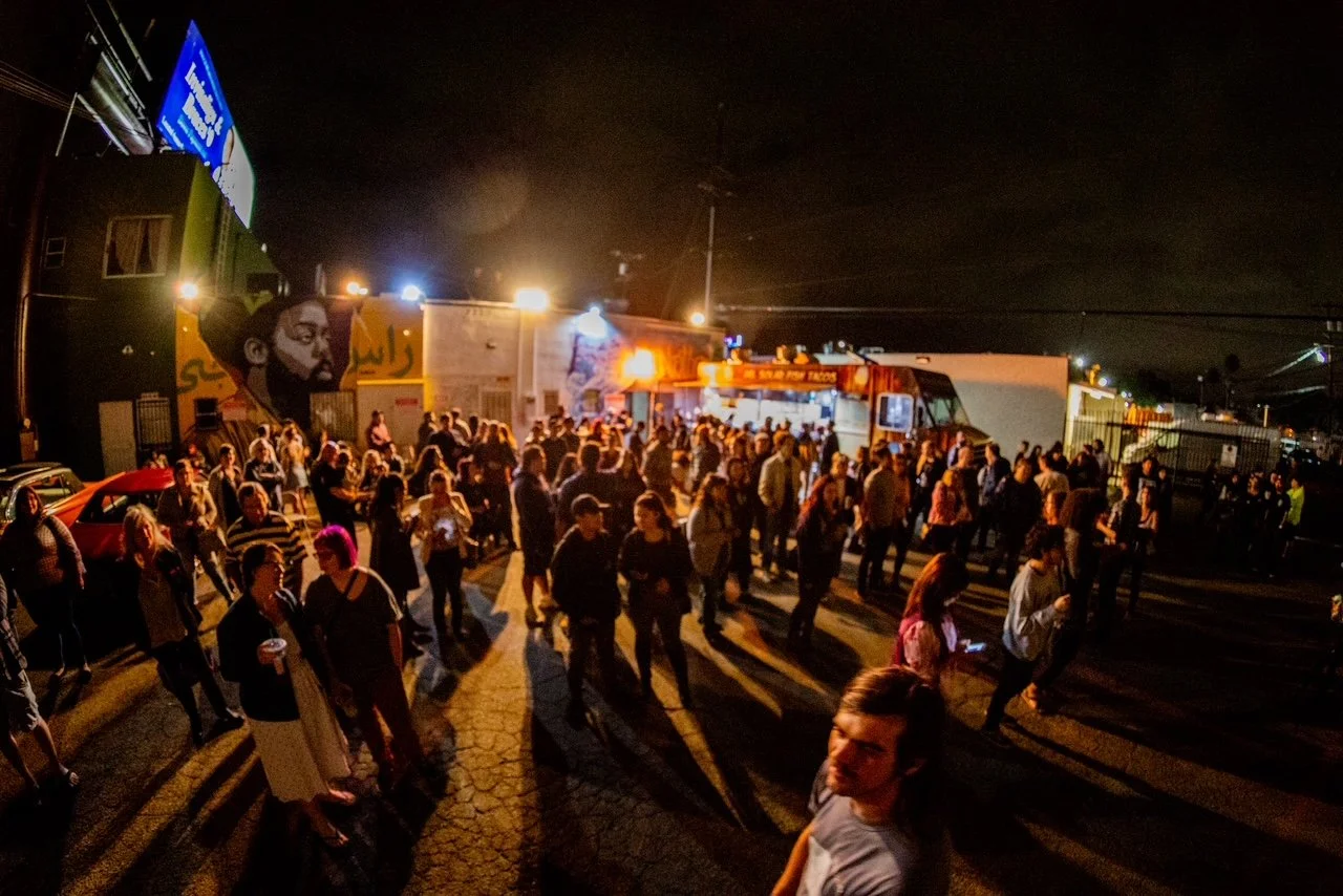 Night scene of a crowded outdoor event with people line dancing, food trucks, and group socializing.