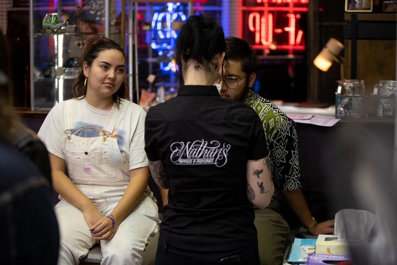A group of young people sitting at a counter in a neon-lit restaurant or bar, with one woman in overalls, a man in glasses, and a woman with tattoos and a black shirt with embroidered text.