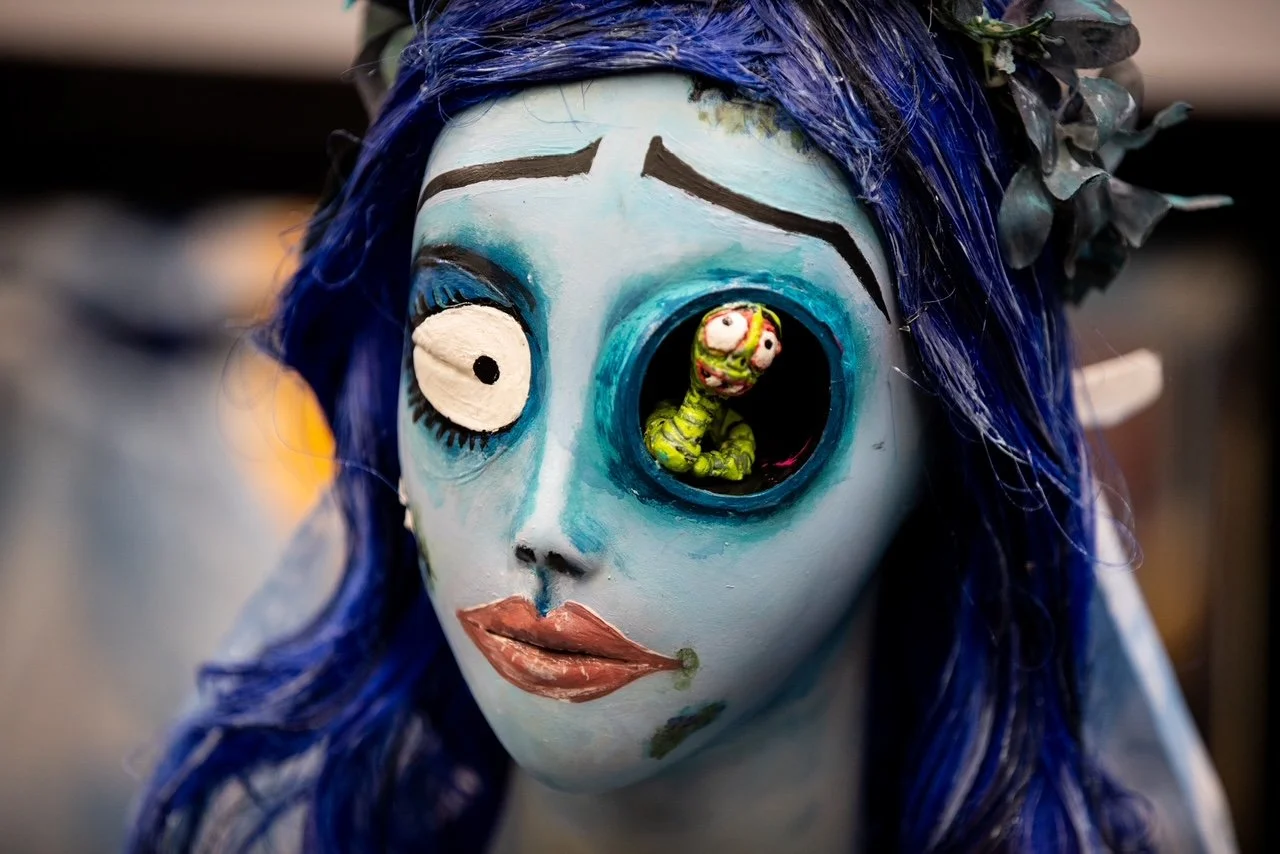 A mask of a woman with blue hair and painted face, with one eye replaced by a circular opening showing a toy caterpillar inside the mask.