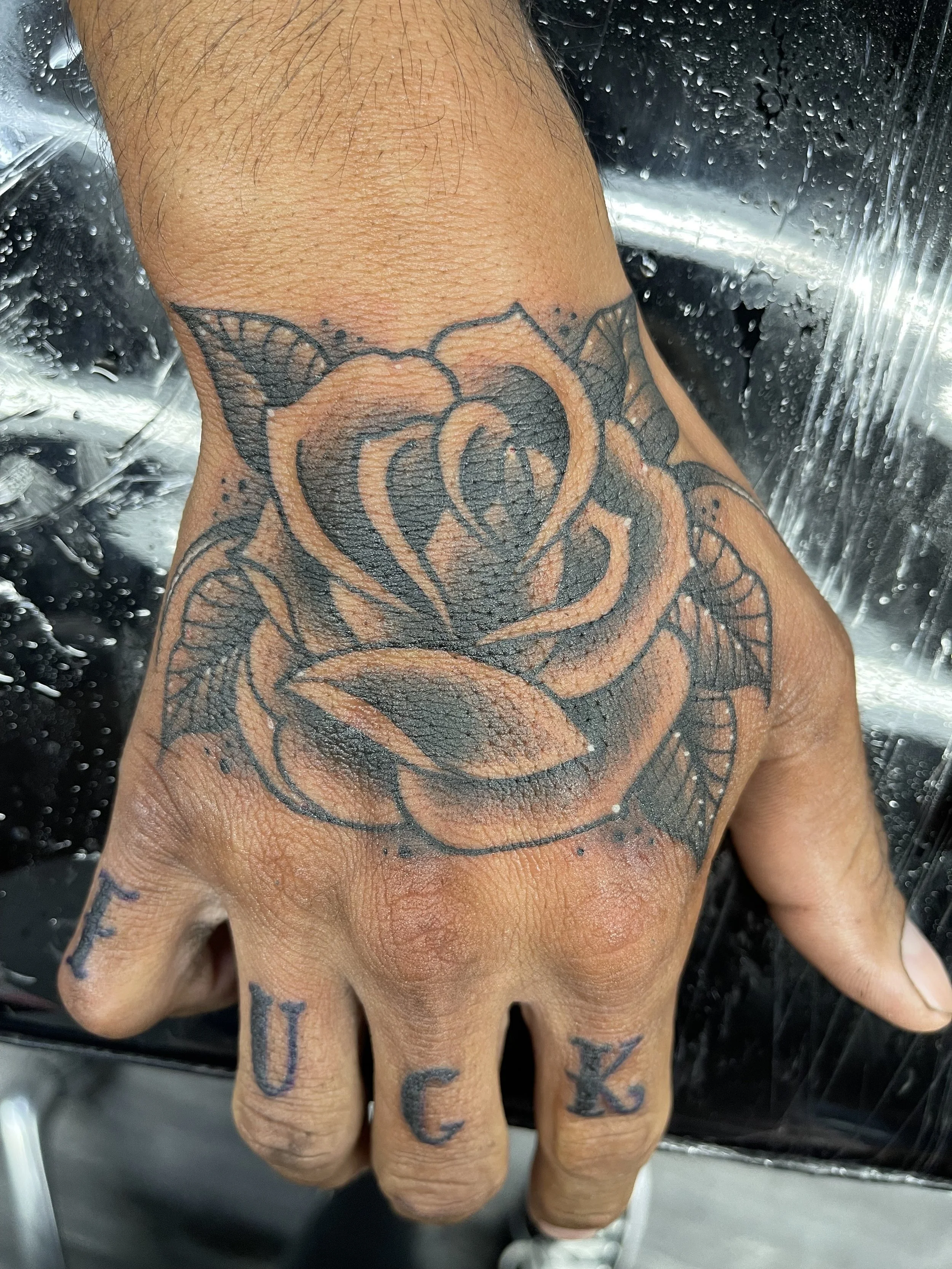 Close-up of a person's hand with tattoos on the fingers and back of the hand, positioned near a shower with water droplets on a glass surface.