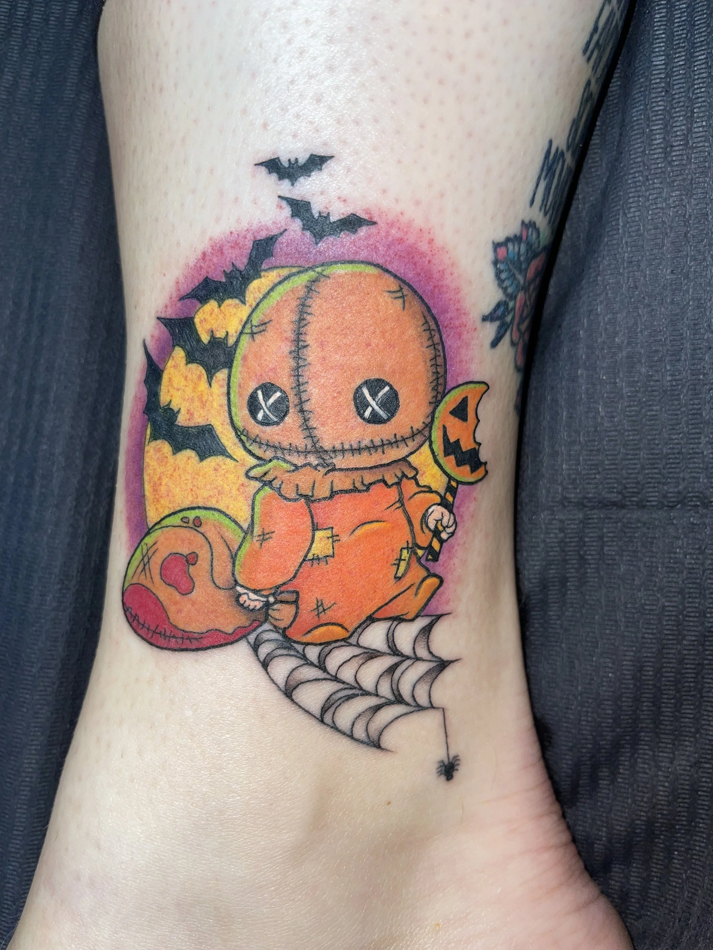 Tattoo of a cartoon pumpkin character with stitched eyes, seated on a pumpkin, surrounded by bats and a spider web at the bottom, with pinkish-red background shading.