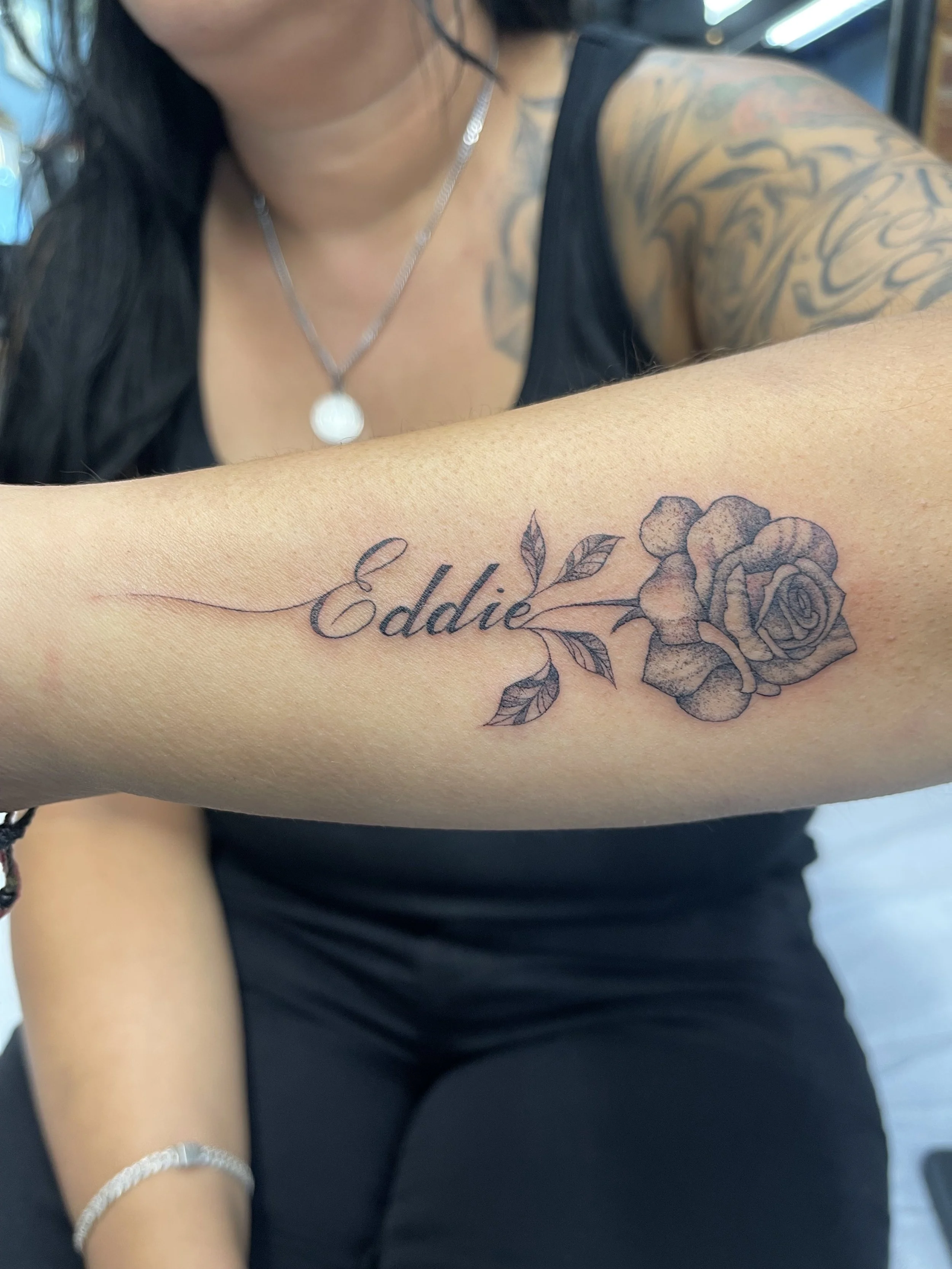 Close-up of a woman with tattoos on her arm, wearing a black sleeveless top, a necklace, and a bracelet, showing a tattoo with the name 'Eddie' and a rose design.