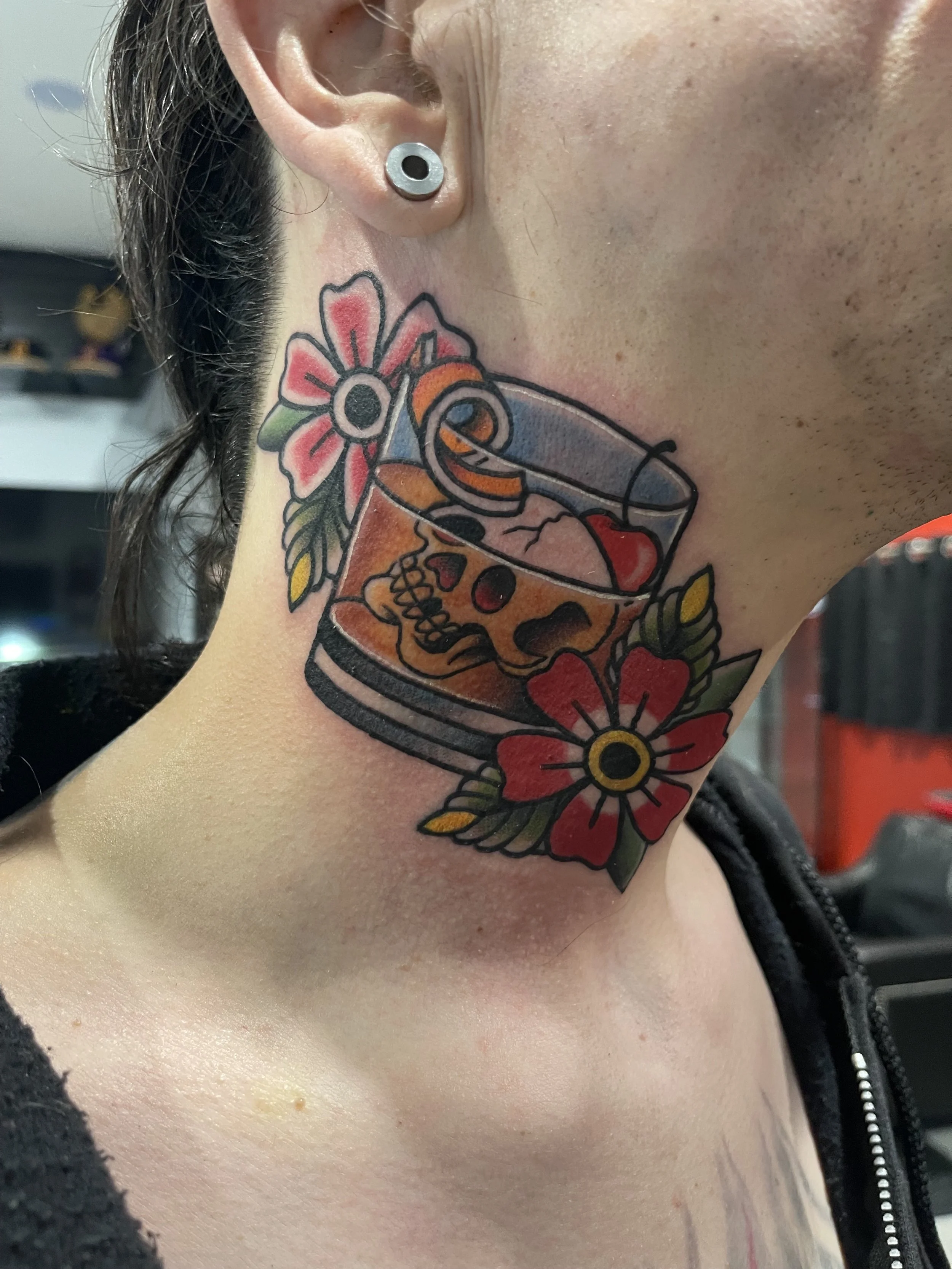 Close-up of a person's neck with a colorful tattoo of a skull within a glass of beverage surrounded by red and pink flowers and green leaves. The person is wearing a black zipper jacket and has a metallic gauge earring.