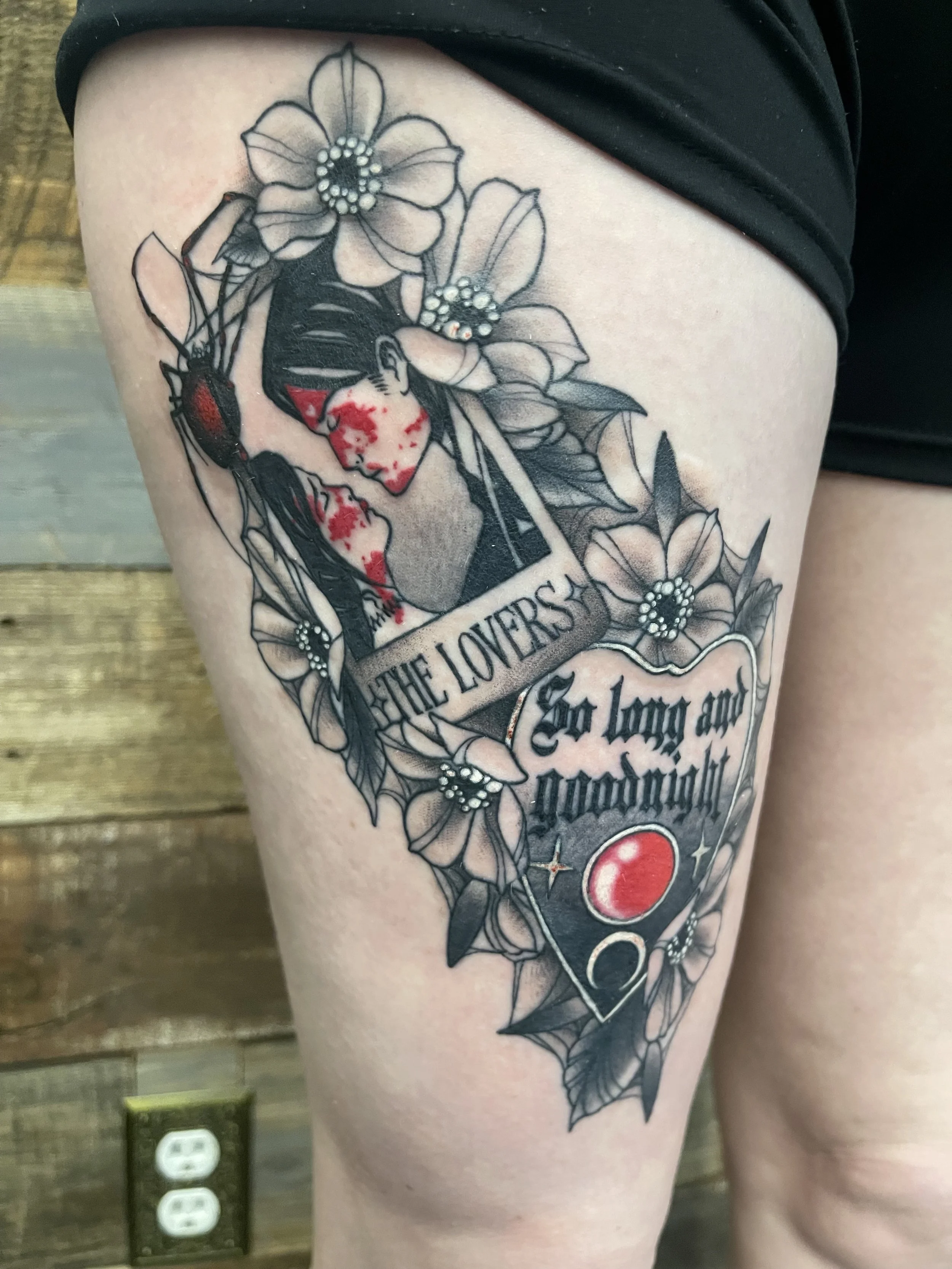 Tattoo of a woman surrounded by flowers and spider webs, with a banner containing text. The woman has blood on her face and hand.
