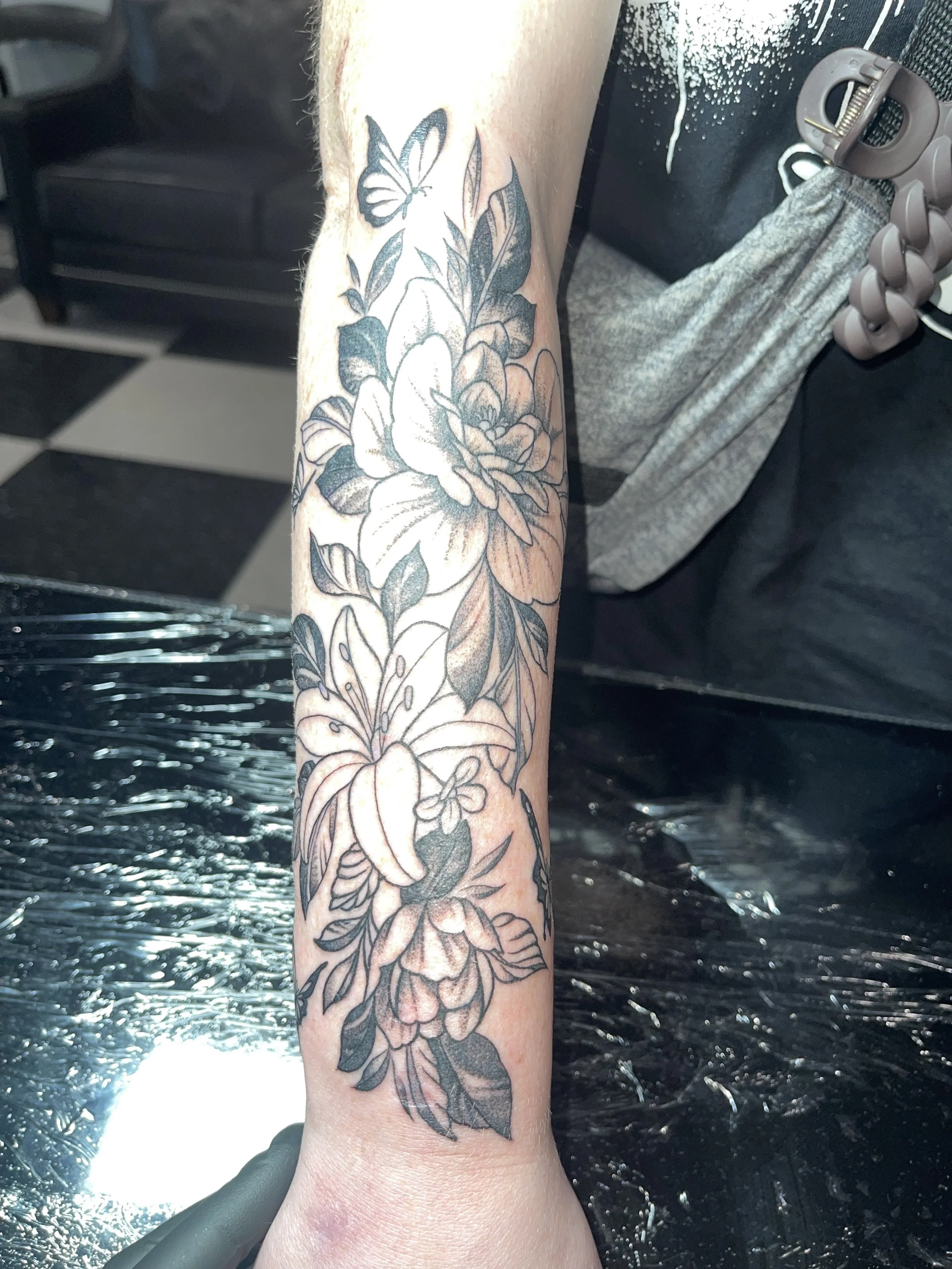 A person's forearm with a black and gray floral tattoo featuring large flowers, leaves, and a small butterfly, being cleaned in a tattoo parlor.