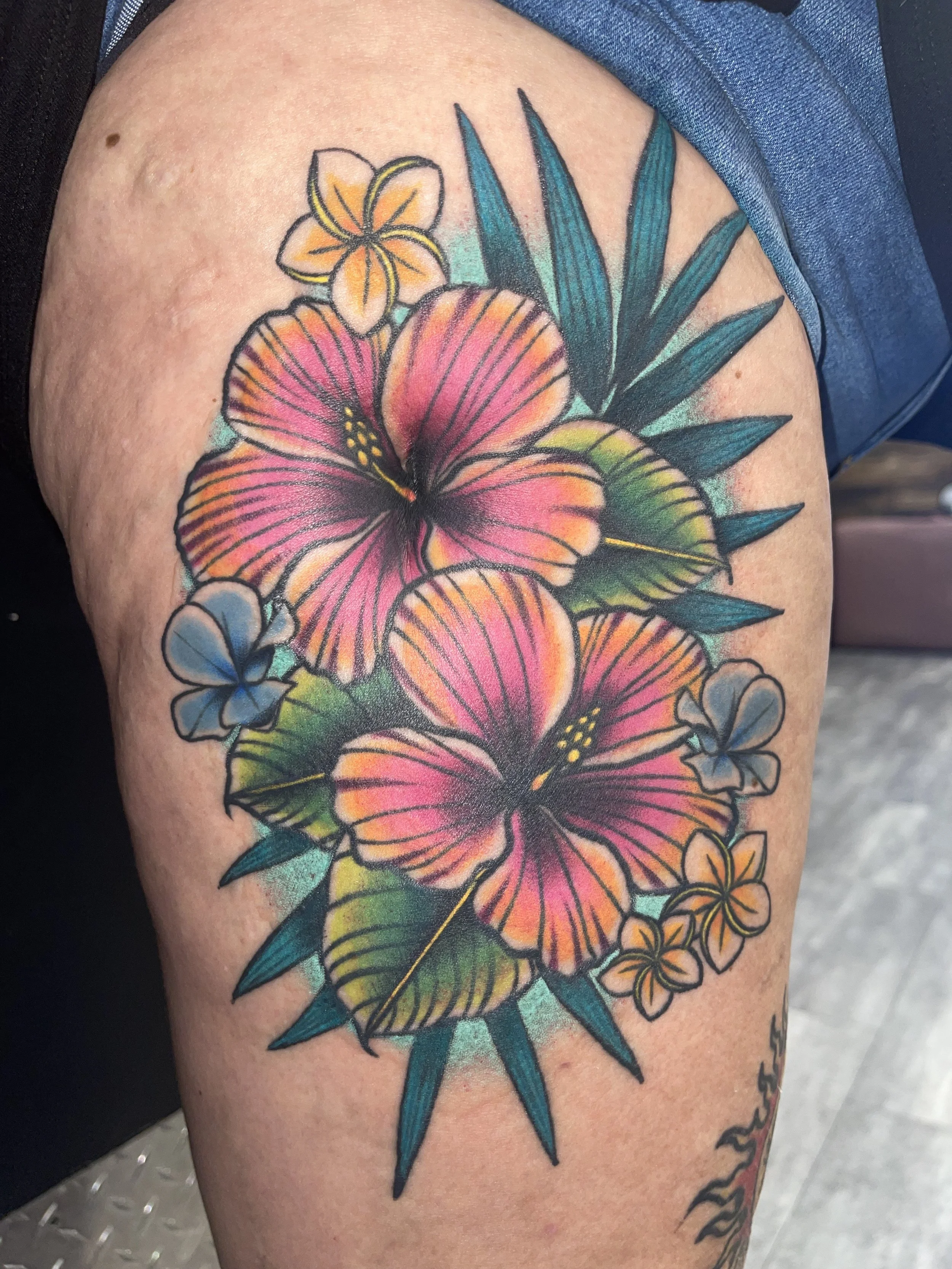 Colorful tattoo of flowers and leaves on a person's upper arm, featuring prominent pink and orange blossoms with green and blue leaves.