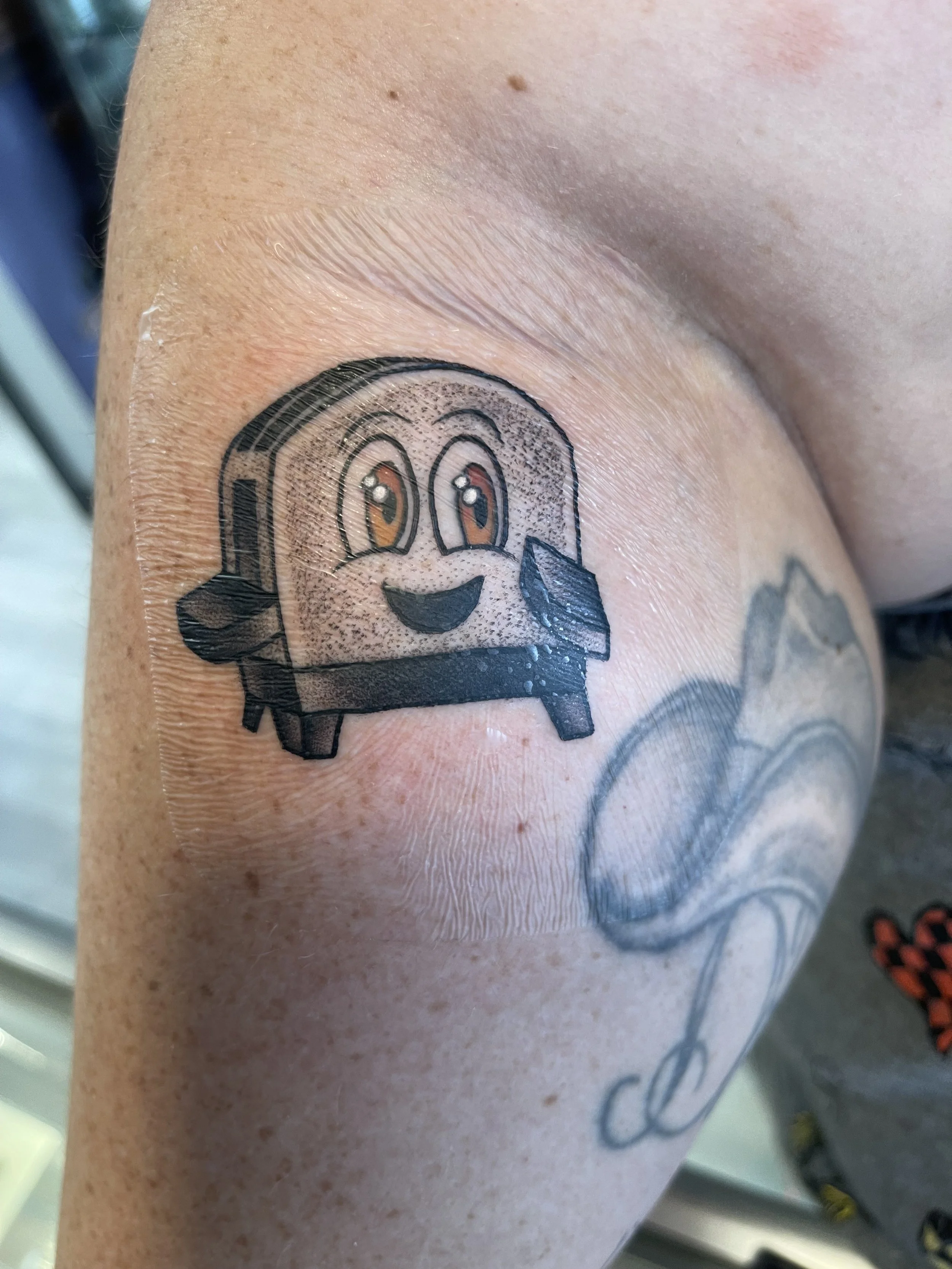 Close-up of a tattoo on skin, featuring a cartoonish smiling face with brown eyes, black outline, and a black hat, with part of a word "amia" visible.