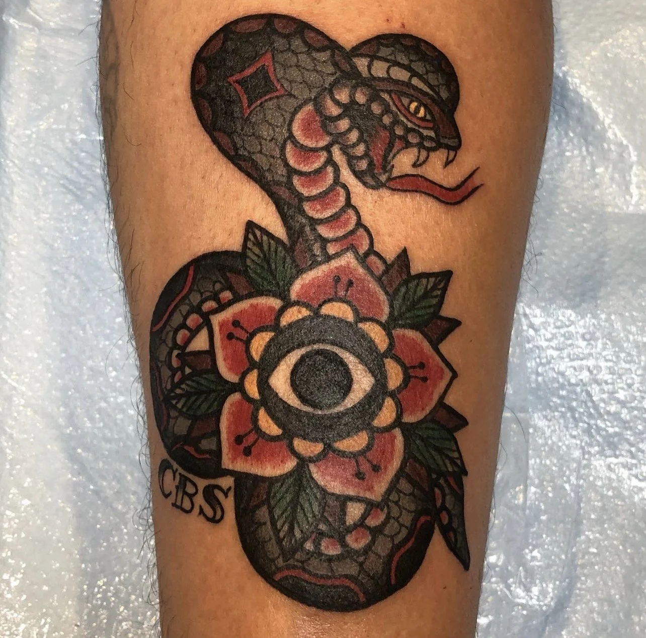 A tattoo of a snake with its head raised, intertwined with a flower that has a central eye and the initials 'CBS' below.