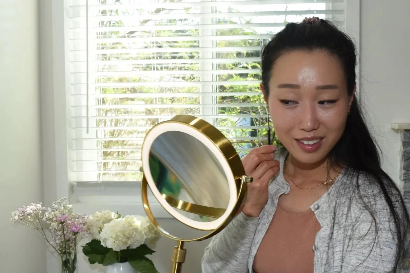 A woman with dark hair in a ponytail is looking into a small, round mirror with a gold frame, adjusting her earrings. She is standing in front of a window with white blinds, with a vase of white and pink flowers on a table nearby.
