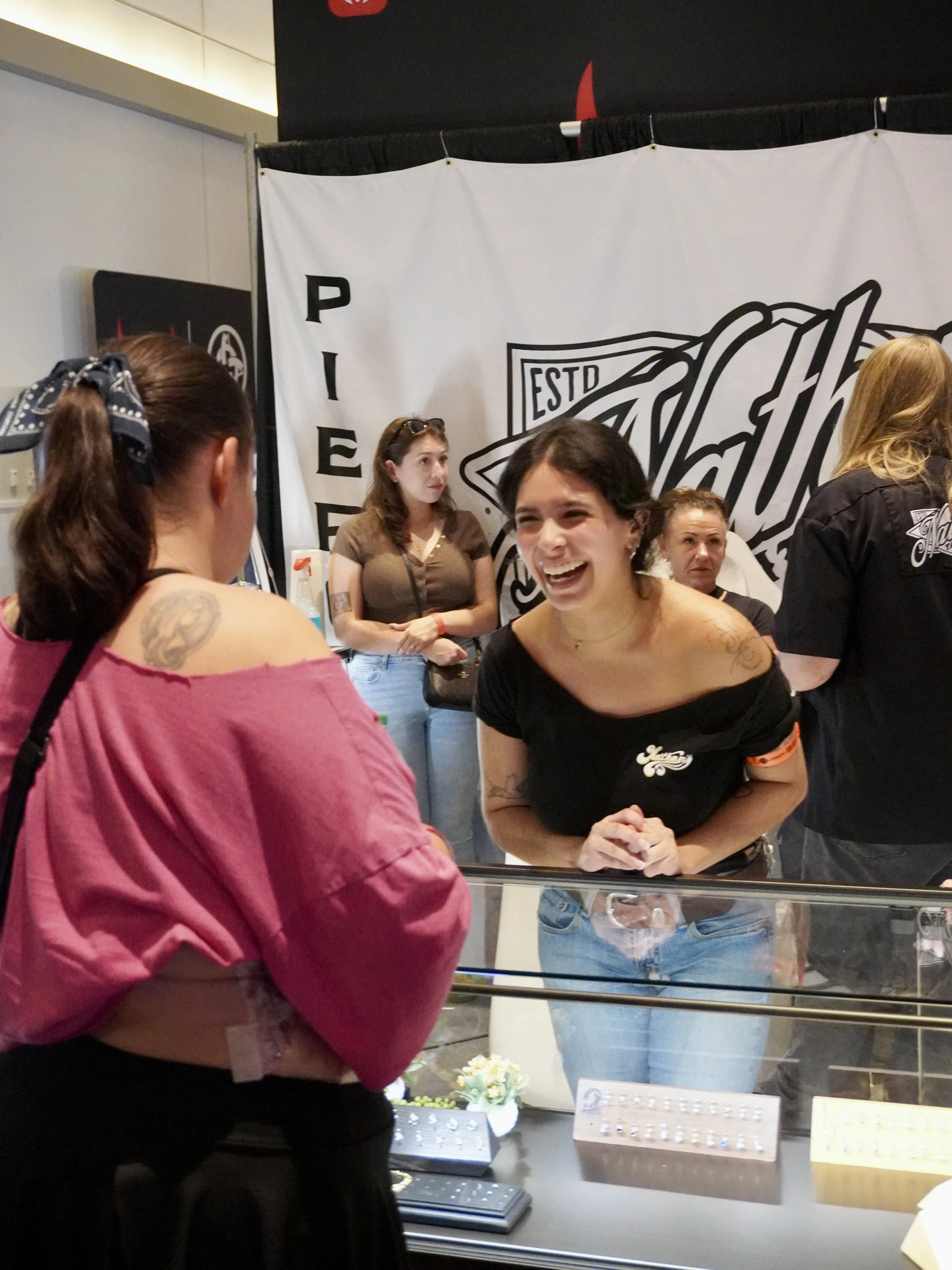 Women shopping at a jewelry store, looking at rings displayed in a case, with a large branded banner in the background.