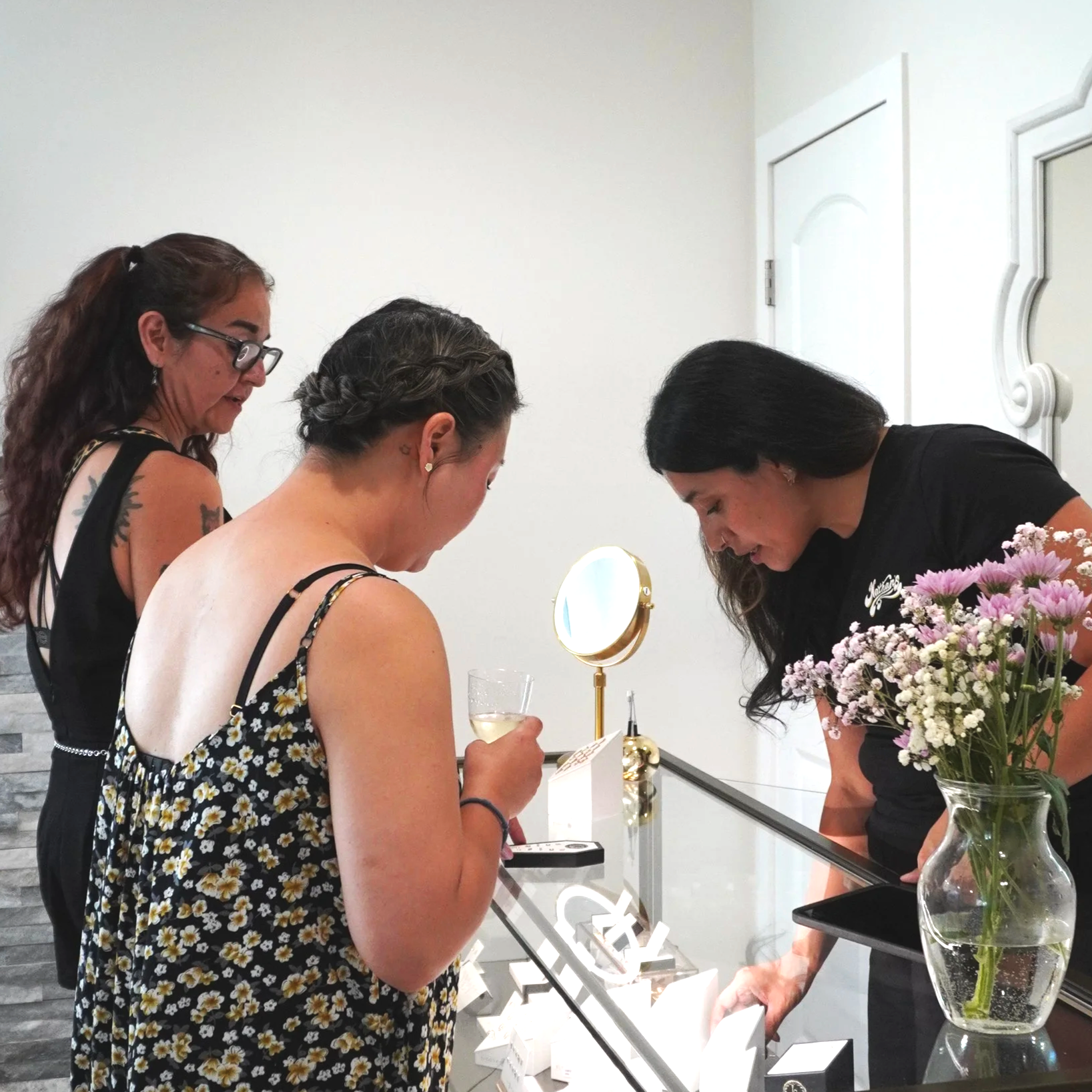 Four women are gathered around a jewelry display case at a store or event, with one woman holding a glass of white wine. A vase of pink and white flowers is on the right, and a small round mirror with a gold frame is on the display.
