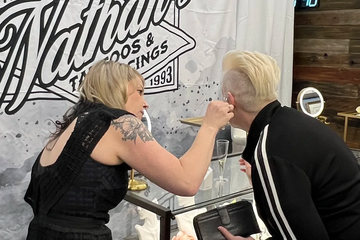 A woman with blonde hair and a tattoo on her arm is applying makeup to a blonde-haired man with a mohawk hairstyle. They are in front of a wall with a large sign that reads 'Nathalie's Tattoos & Piercings' and appear to be at a beauty or tattoo boutique with makeup mirrors and champagne glasses on the table.