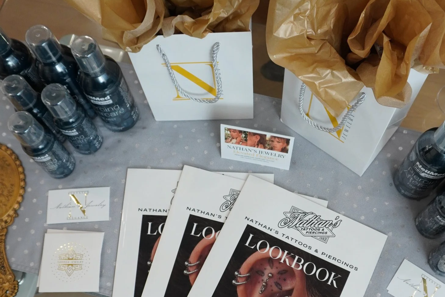 Display table with promotional posters for Nathan's Tattoos & Piercings, bottles of a product labeled 'Healing Care,' paper bags with tissue paper, and a small business card reading 'Nathan's Jewelry.'