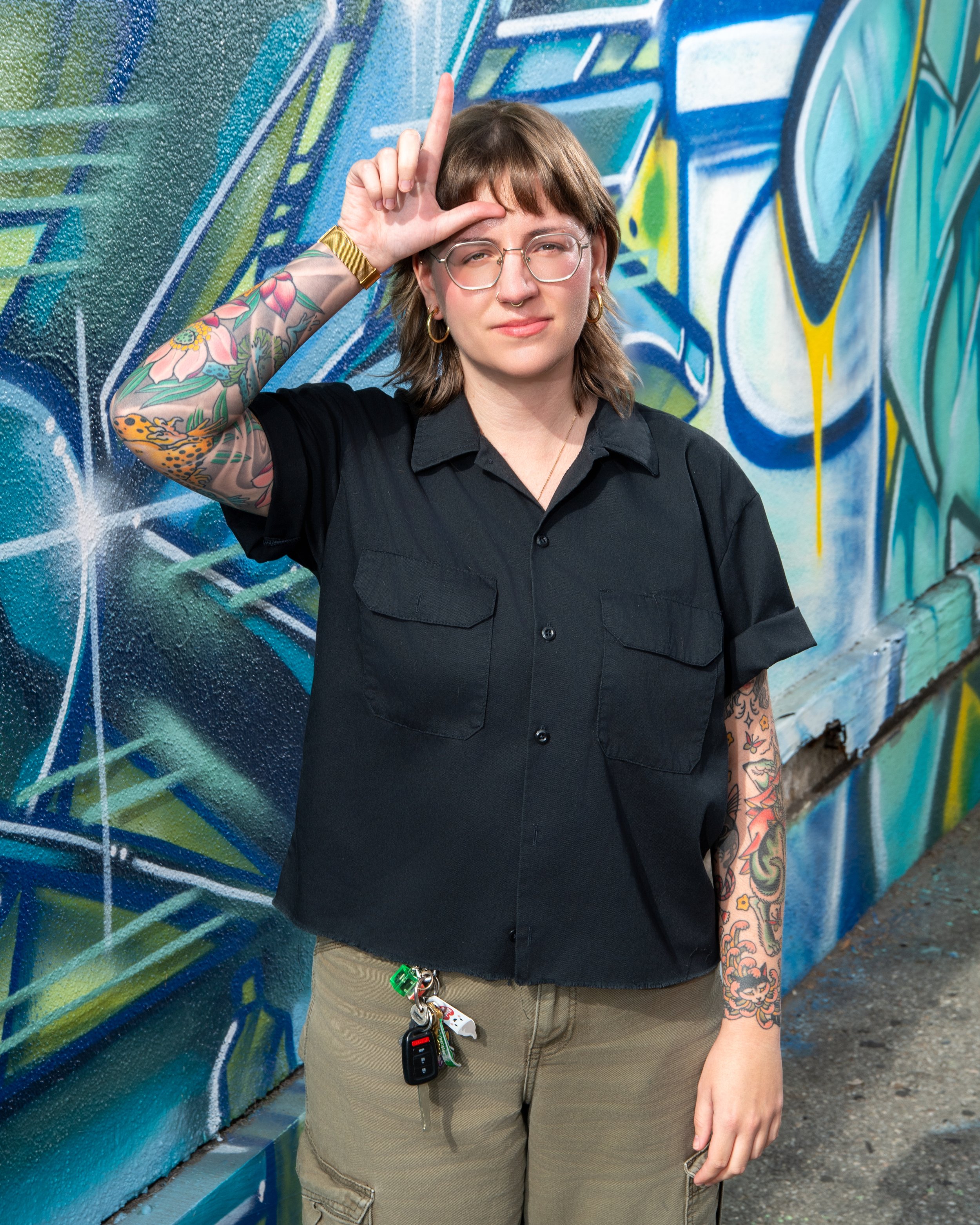 A person with glasses, tattoos, and wearing a black shirt and khaki pants, standing in front of a colorful graffiti wall, making an 'L' shape with their hand on their forehead.