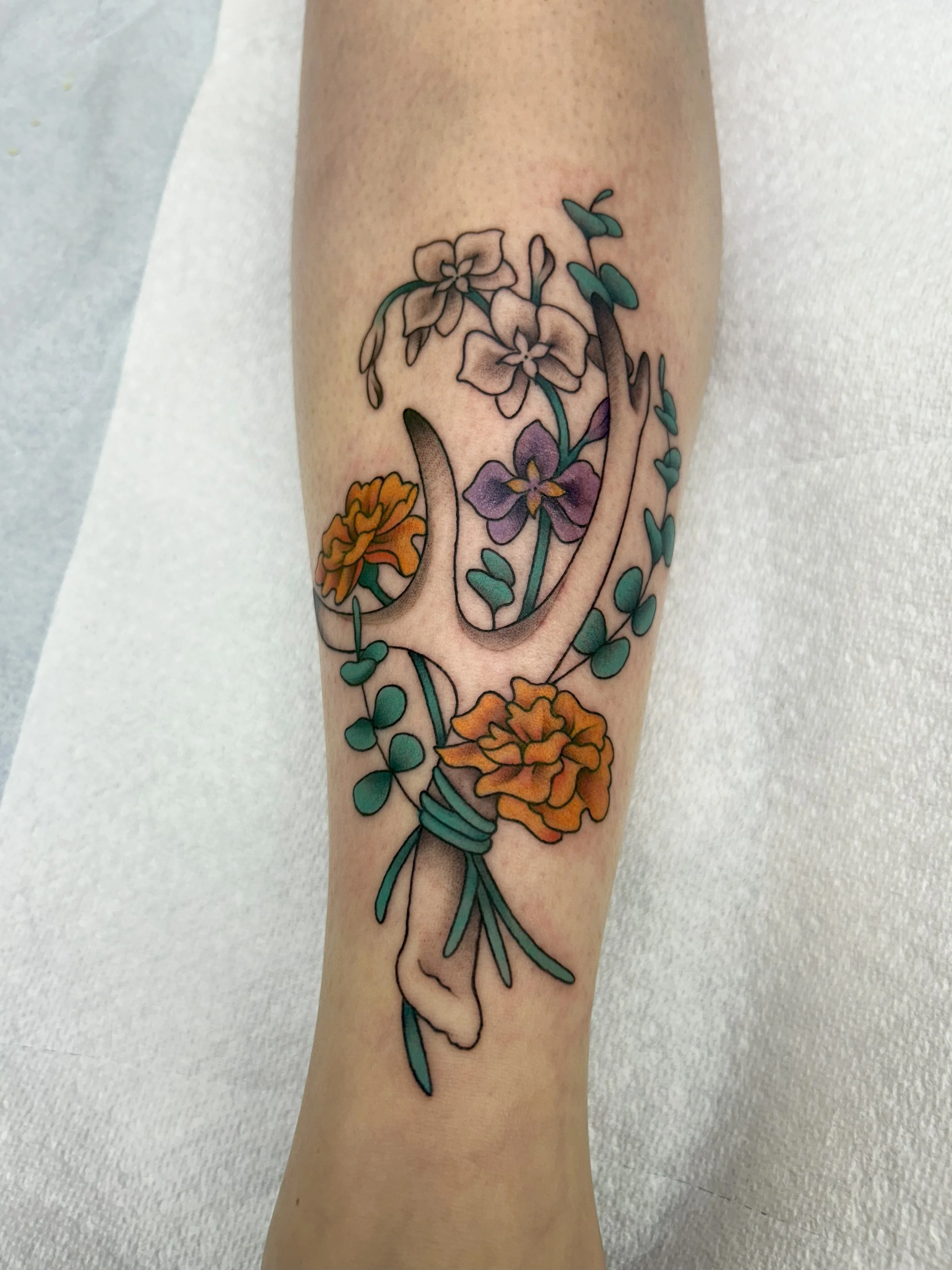 A tattoo of a bouquet of colorful flowers with a daffodil, pansy, and other blossoms, tied together with a ribbon, on a person's forearm.