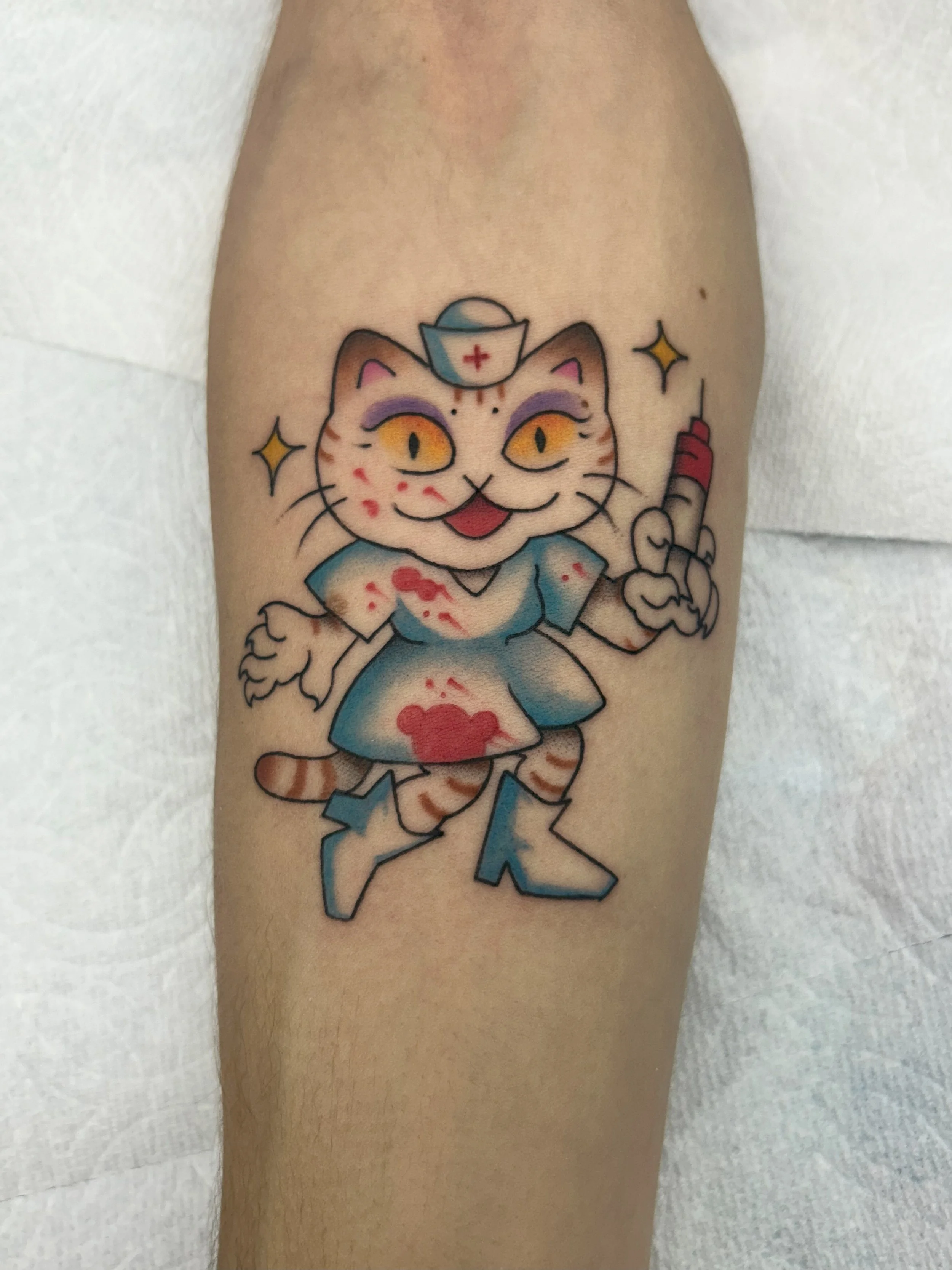 Colorful tattoo of a smiling anthropomorphic cat dressed as a nurse or doctor, holding a syringe, with a nurse's cap and medical symbols, on a person's upper arm.