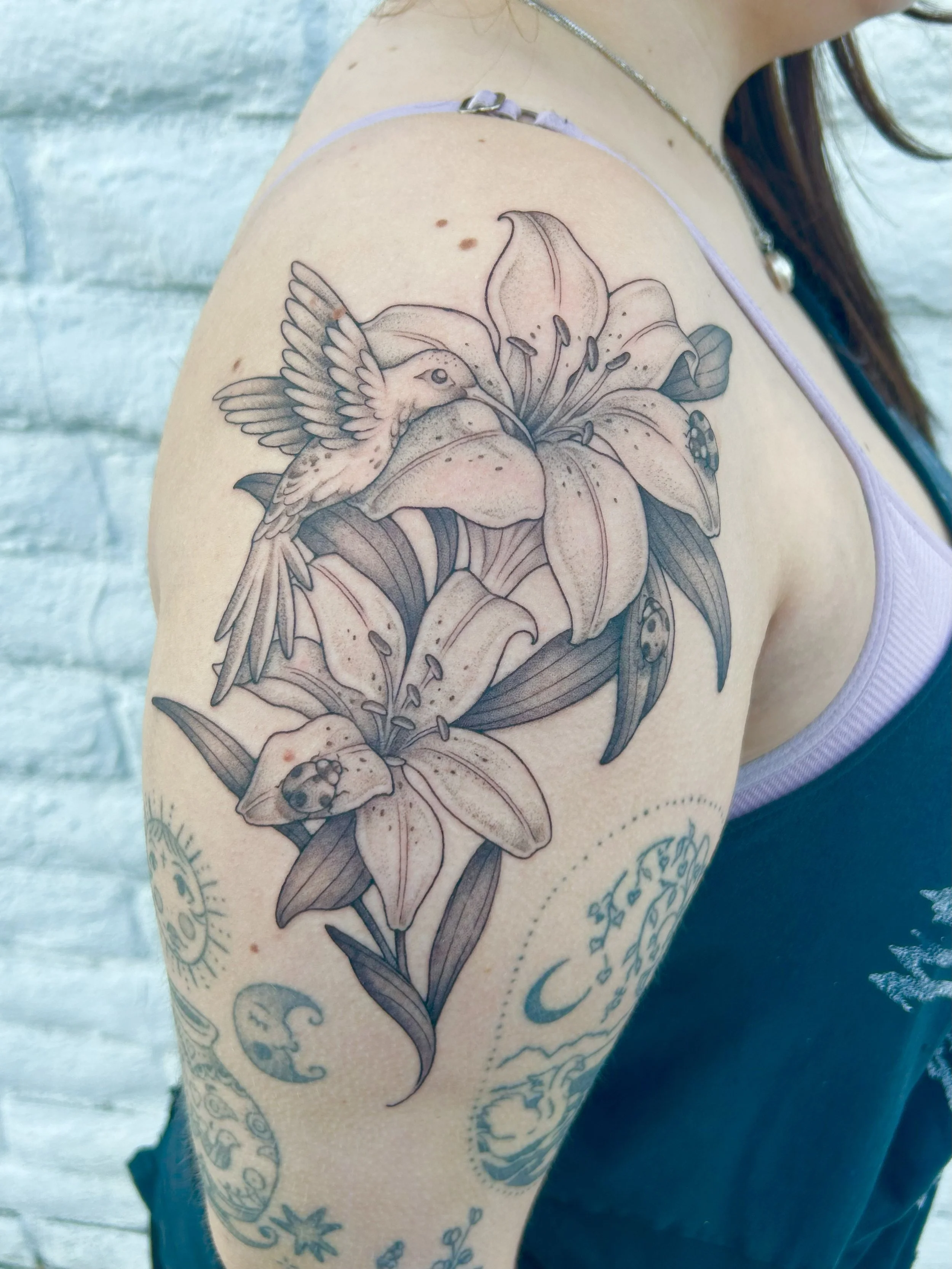 A close-up of a person's upper arm and shoulder showing a detailed tattoo of a hummingbird flying near a large lily flower with leaves.
