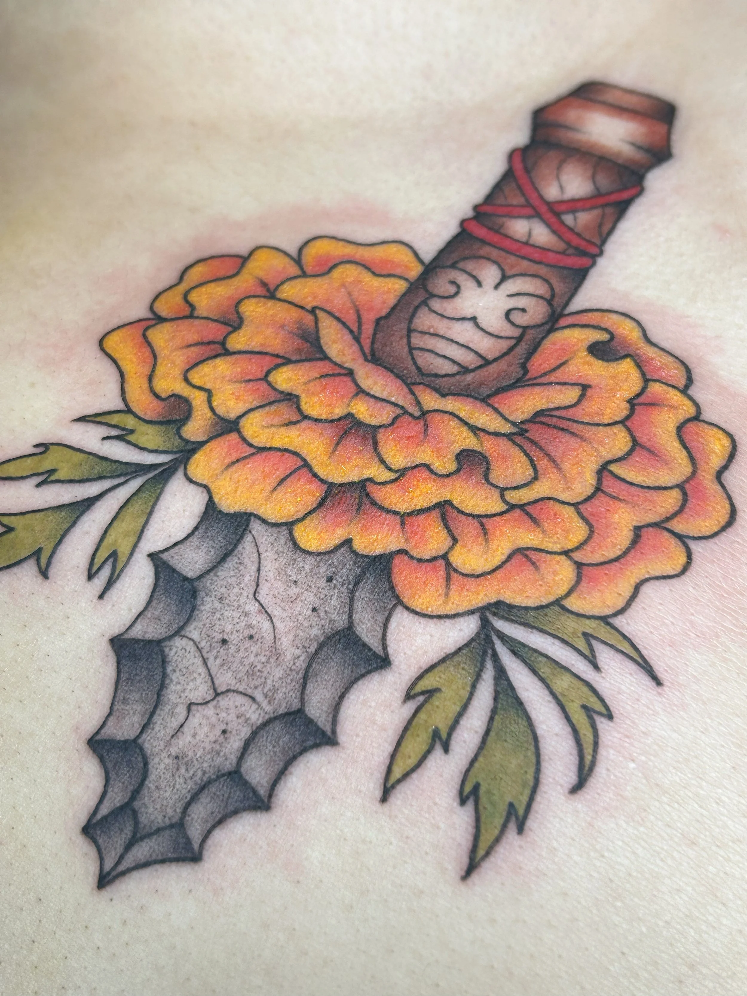 Tattoo of a flower with orange and yellow petals, green leaves, and a dagger piercing through the flower.