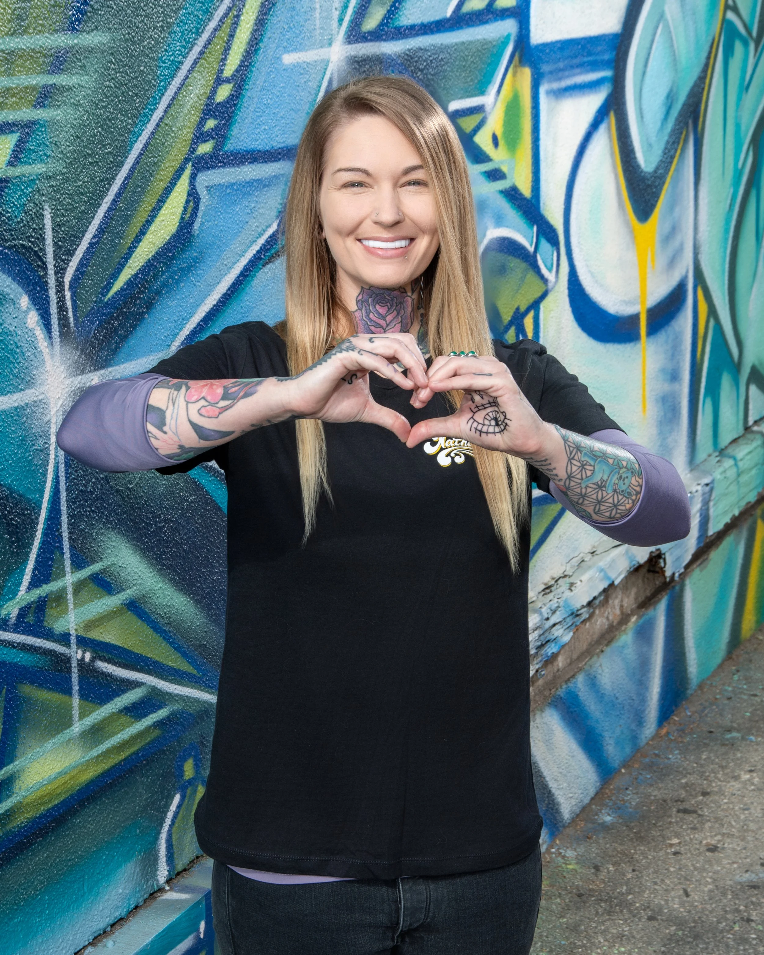 A smiling young woman with tattoos on her arms and neck forming a heart shape with her hands, standing in front of a colorful graffiti wall.