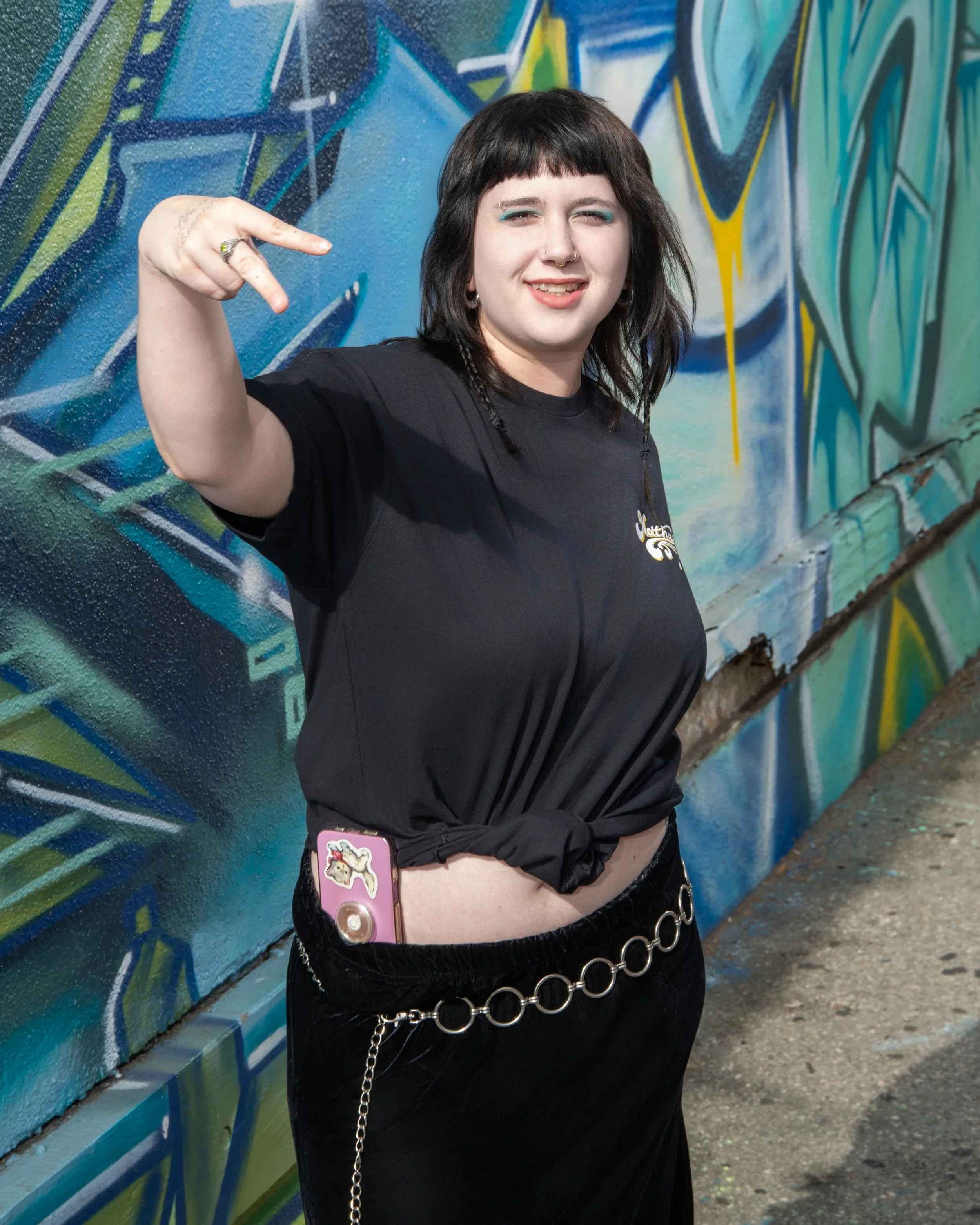 A person with black hair, wearing black clothing with silver chains, standing in front of a colorful graffiti wall, making a peace sign with their hand.