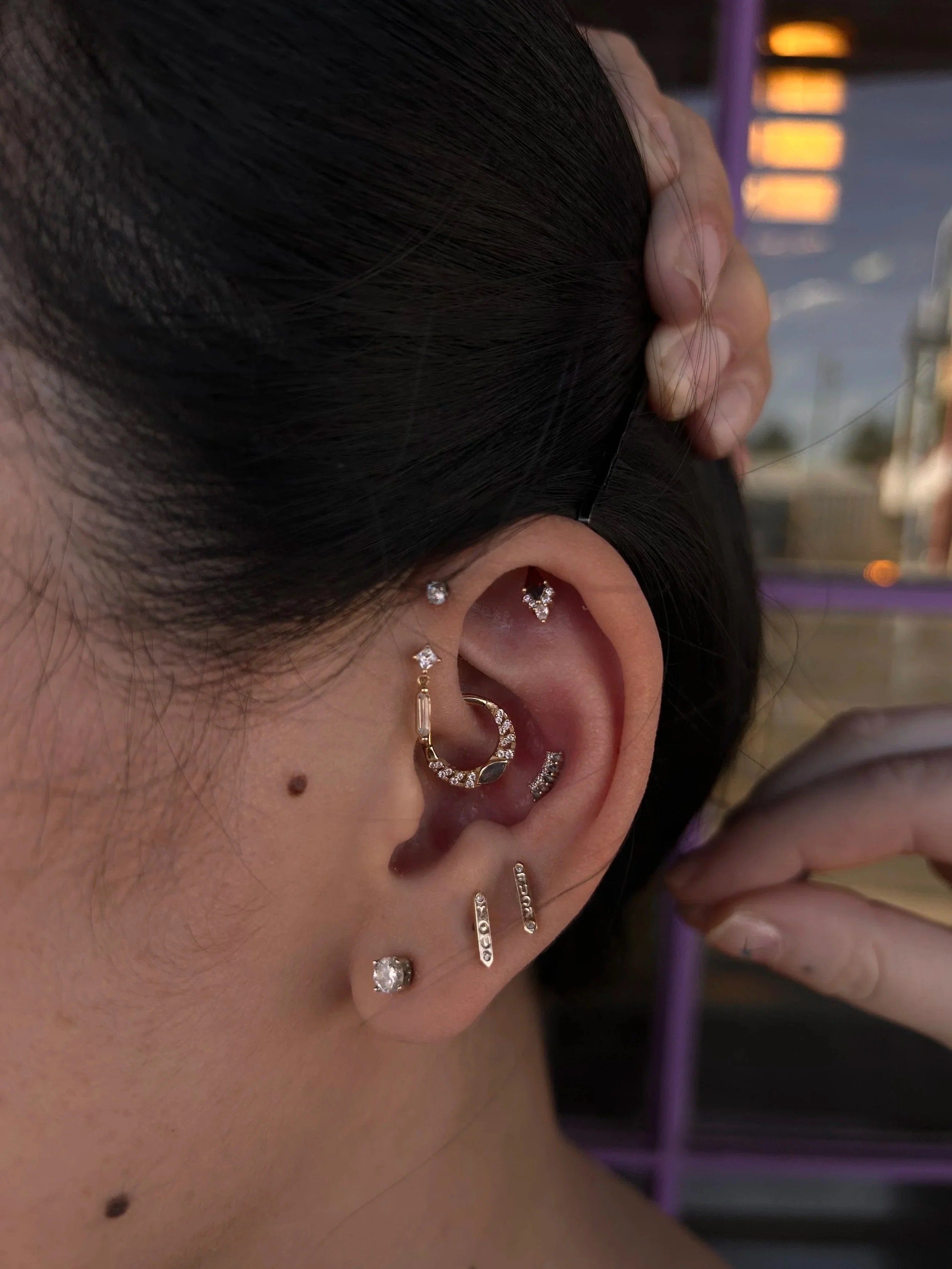 Close-up of a person's ear adorned with multiple earrings, including studs and hoops, with dark hair pulled behind the ear, and a background with warm lighting.