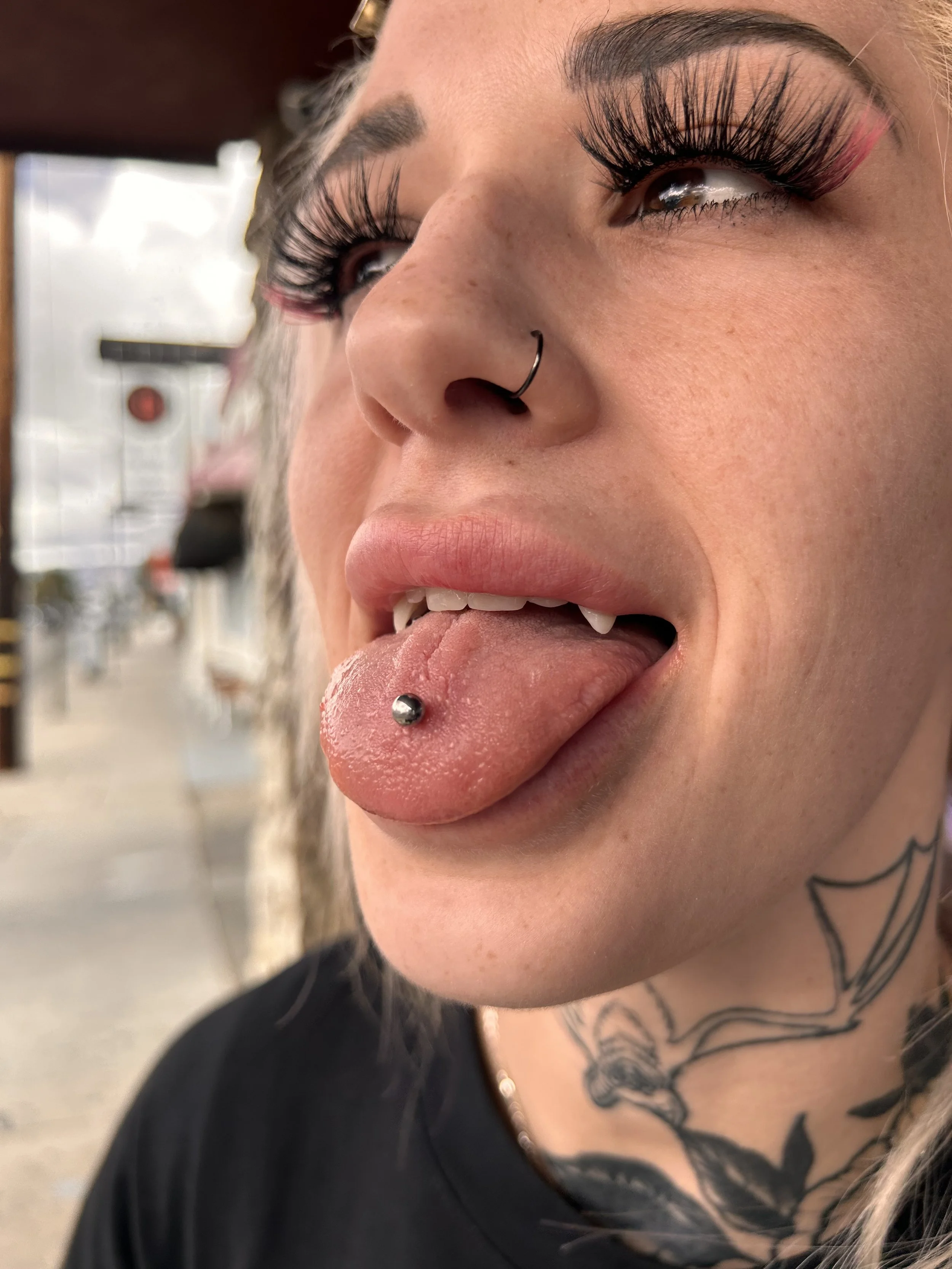 Close-up of a person with thick, long eyelashes, a septum piercing, and a tongue piercing. They have a tattoo on their neck and are wearing a black shirt. The background is indoors and blurred.